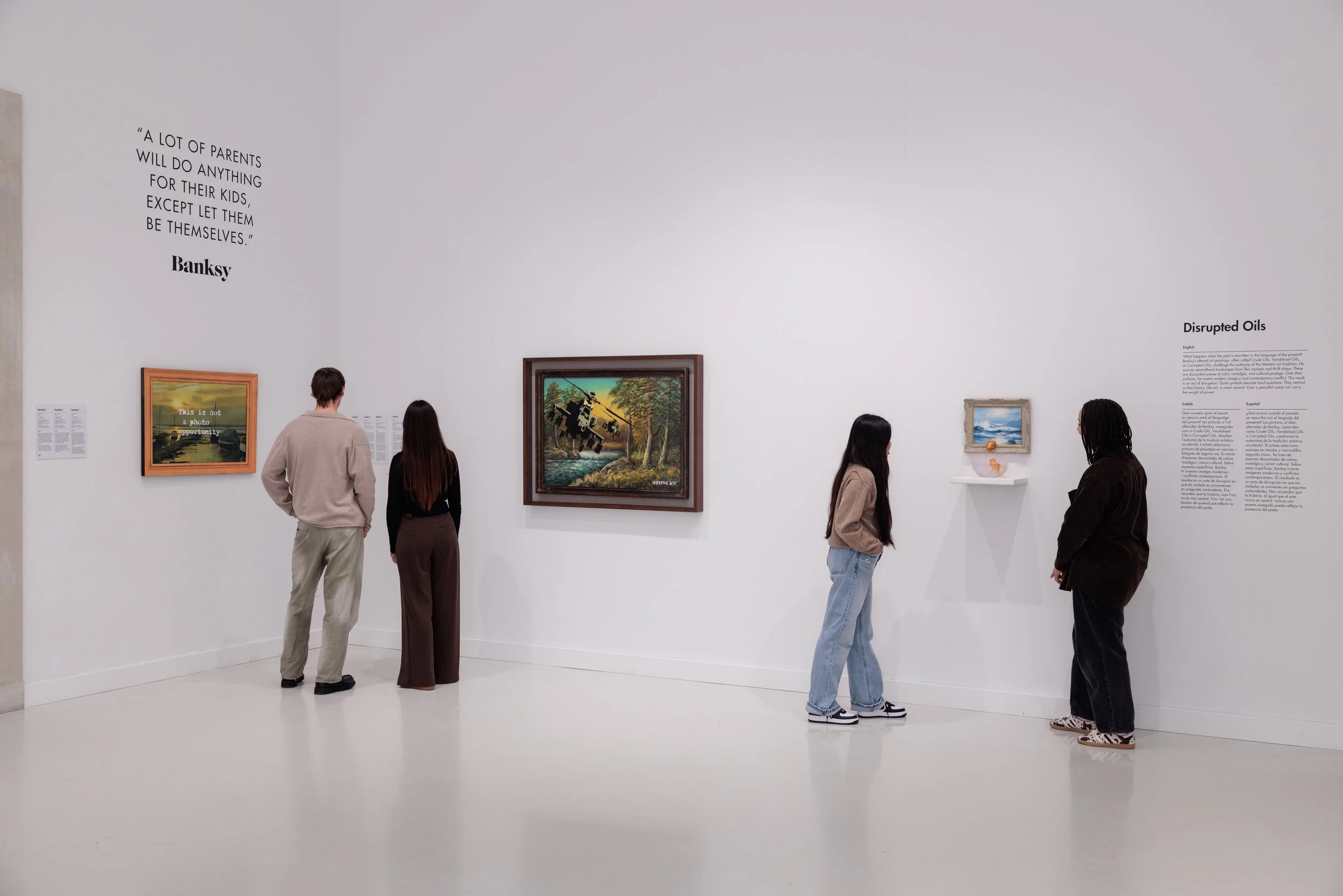 Fünf Menschen betrachten Kunstwerke in einem Museum. Die Wand zeigt ein Zitat von Banksy, und es gibt mehrere Gemälde an der Wand.