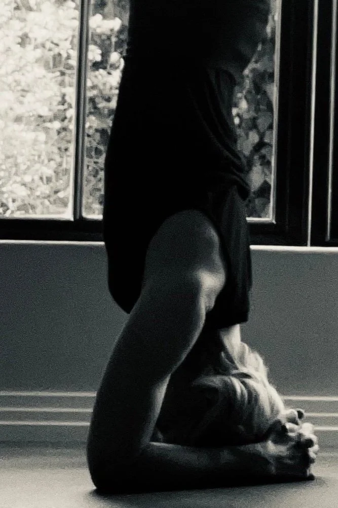 A person practicing yoga or meditation indoors, kneeling on the floor with their hands clasped in front of them, near a window with natural light coming through.
