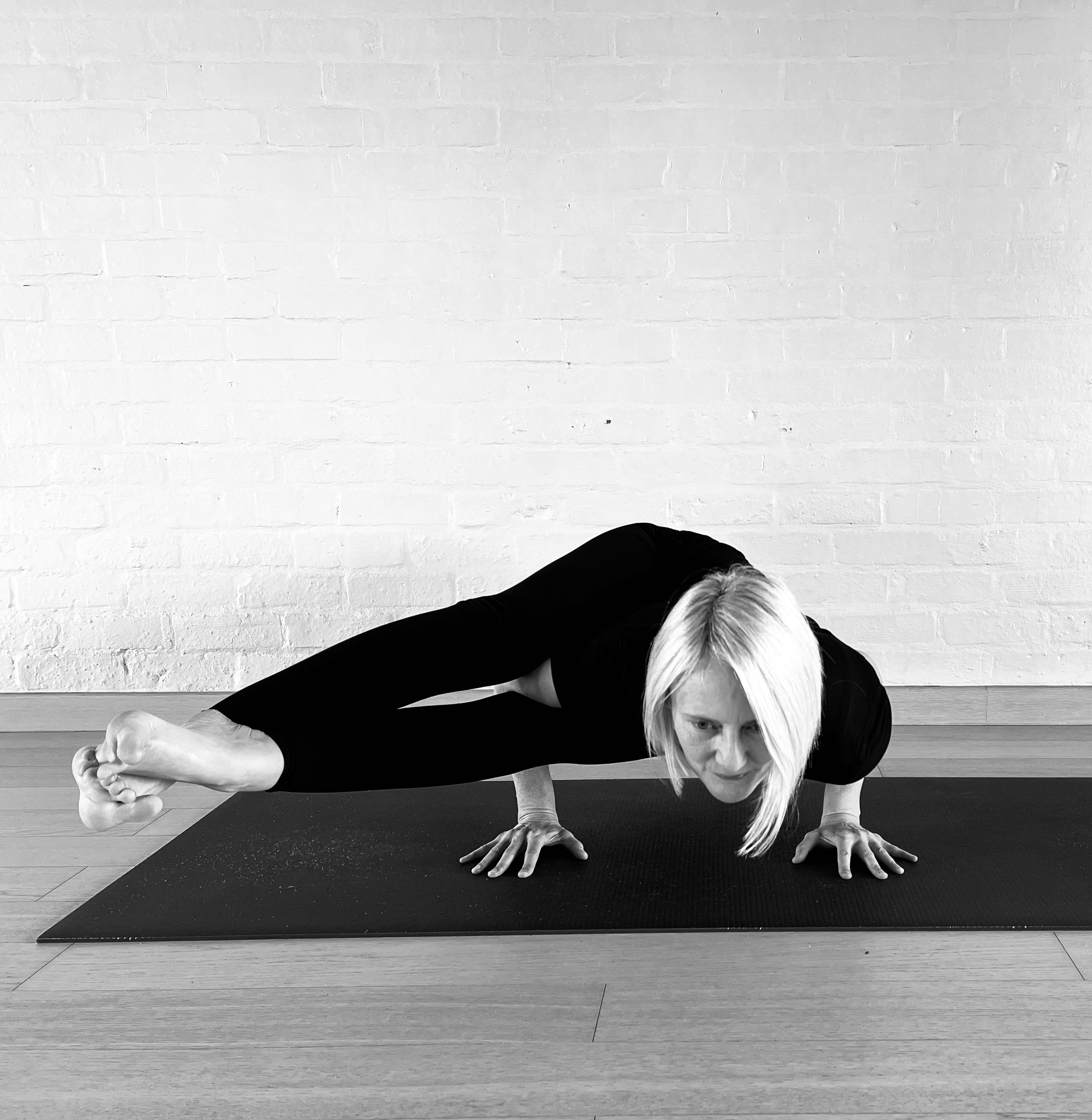 A woman with blonde hair doing astavakrasana on a black mat against a white brick wall. She is supporting herself on hands and feet, with one arm raised and eyes focused forward.