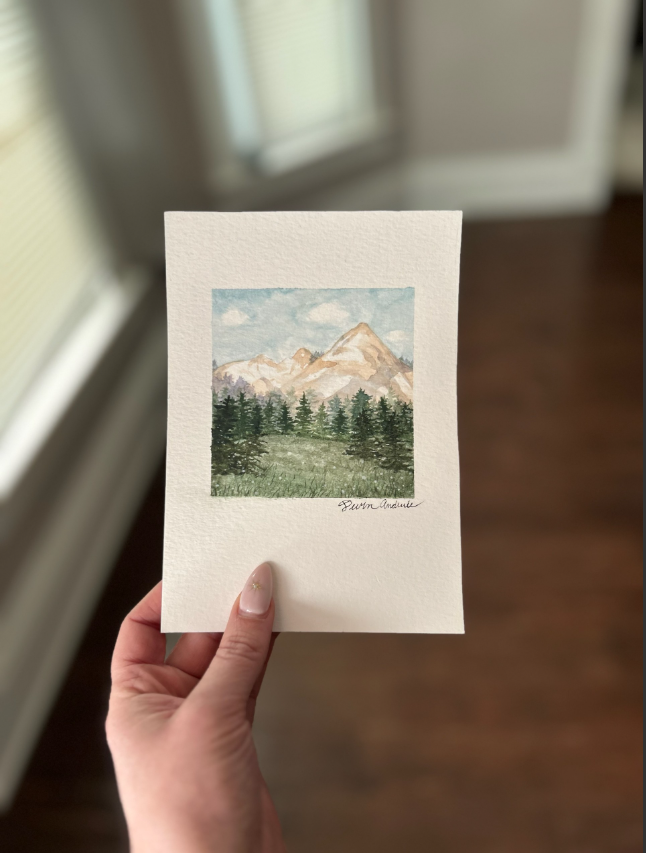 Hand holding a watercolor painting of mountains and trees on white paper.
