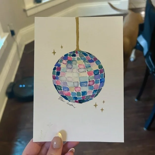 Hand holding a watercolor painting of a disco ball with colorful tiles and golden stars on a white paper.