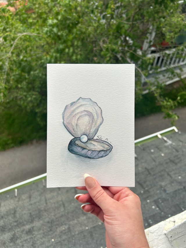 Hand holding a watercolor painting of an open oyster shell with a pearl inside, against a blurred outdoor background.