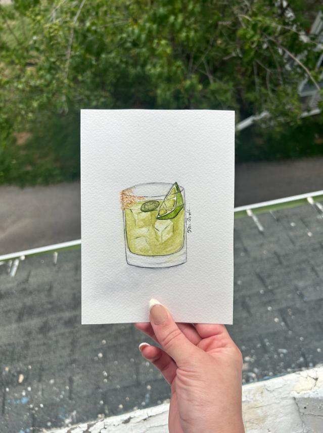 Hand holding a watercolor painting of a green cocktail in a glass, garnished with lime and salt, against an outdoor background.