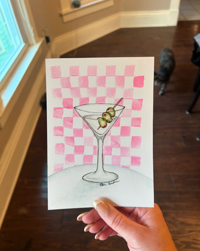 Hand holding a watercolor painting of a martini glass with three olives on a stick, set against a pink checkerboard background.