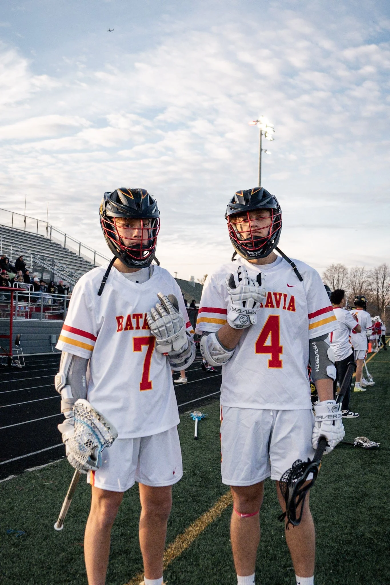 Two young male lacrosse players in white uniforms with red and yellow stripes, black helmets with face cages, and gloves standing on a sports field. The player on the left has the number 7, and the player on the right has the number 4. They are holdi