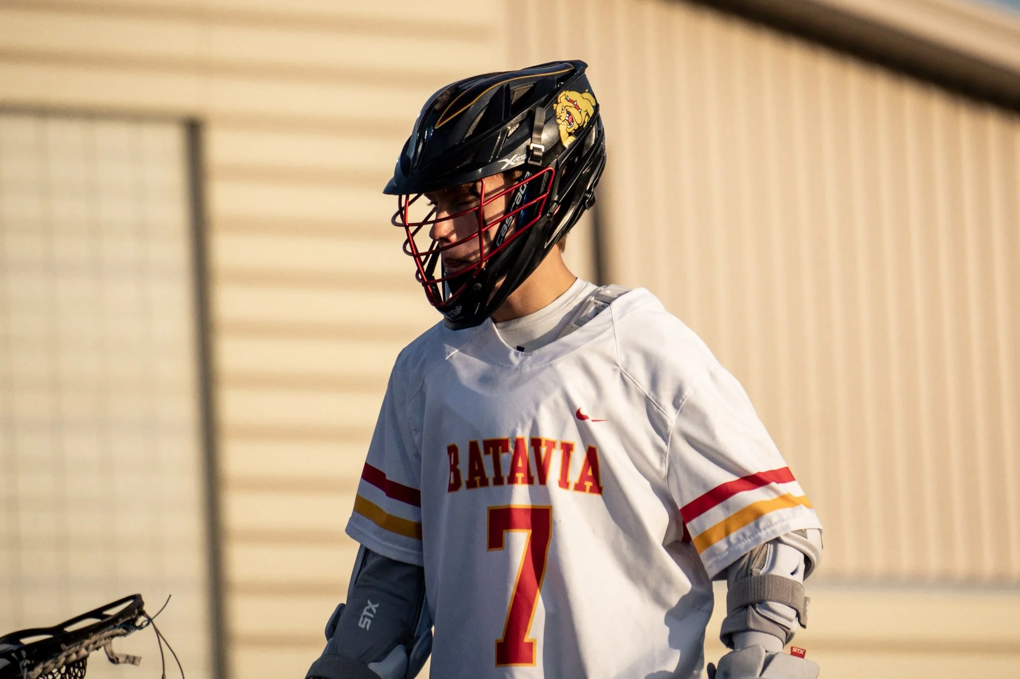 A youth lacrosse goalie wearing a helmet, jersey with number 7, and protective gear stands outdoors with a beige building in the background.