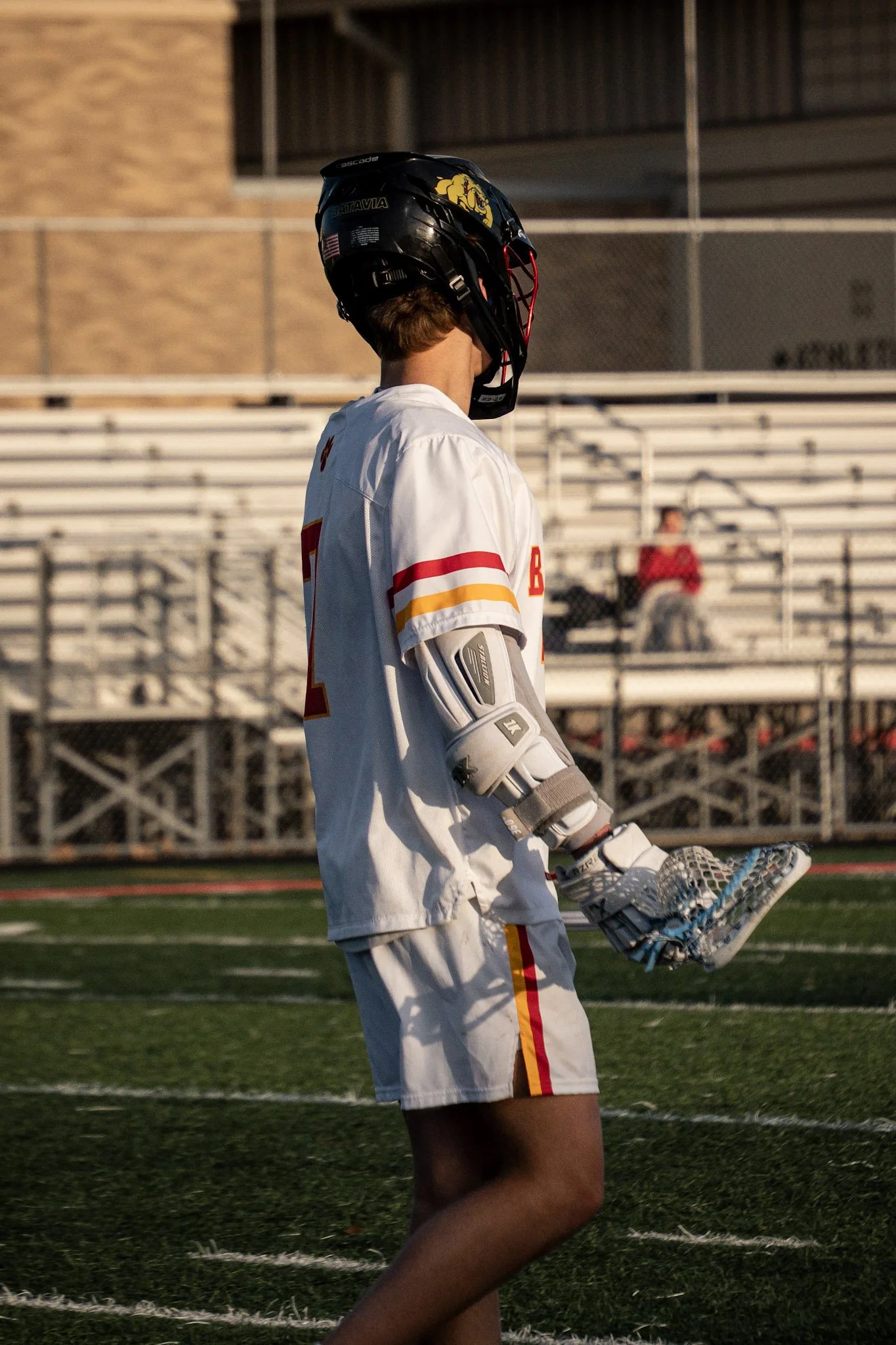 A lacrosse player wearing a helmet, white jersey with red and yellow stripes, and protective gear, standing on a field with empty bleachers in the background.