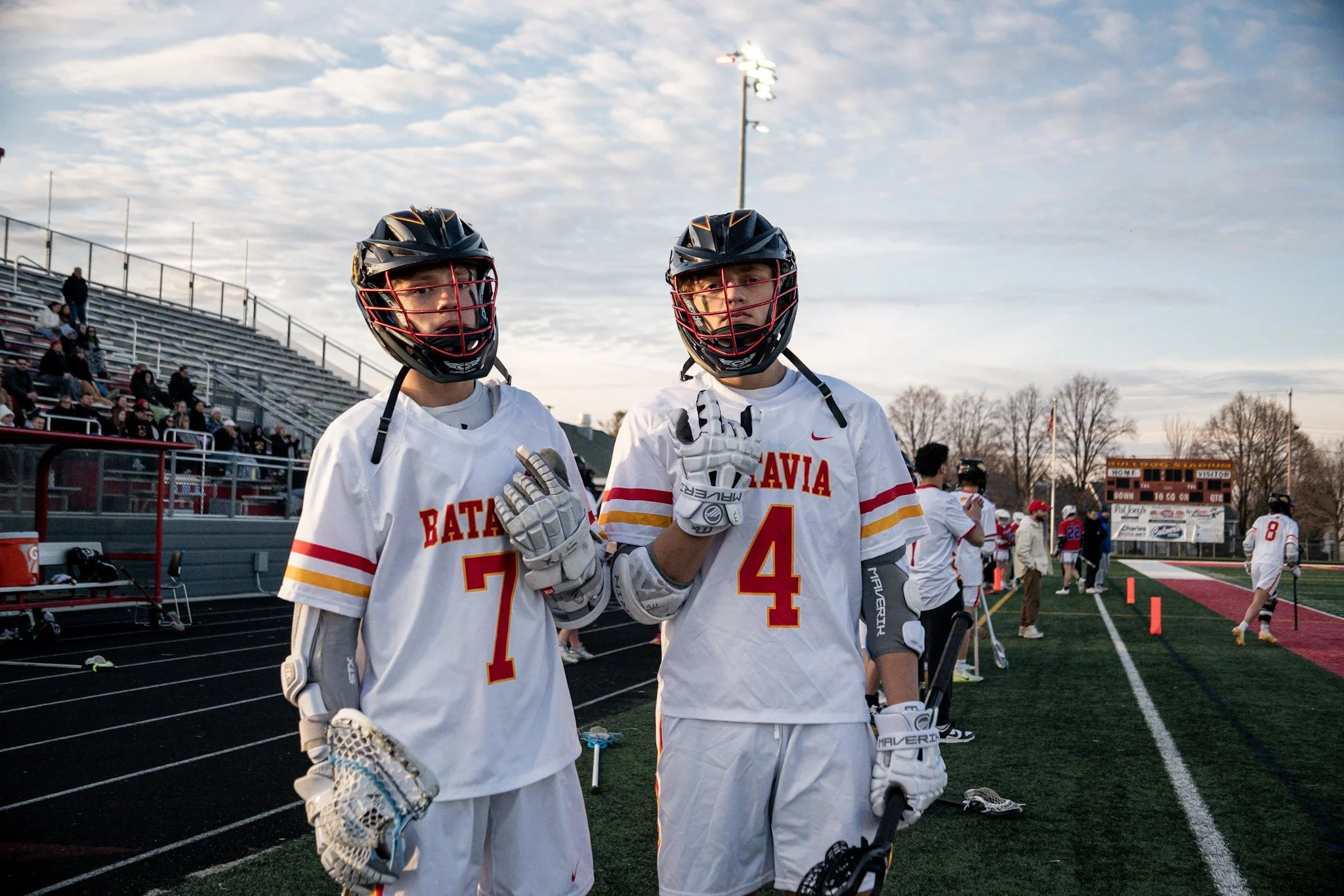 Two male lacrosse players in white uniforms with red and yellow accents, wearing black helmets with red facemasks, standing on a sports field during a game or practice, with a track, spectators in stands, and other players in the background at sunset