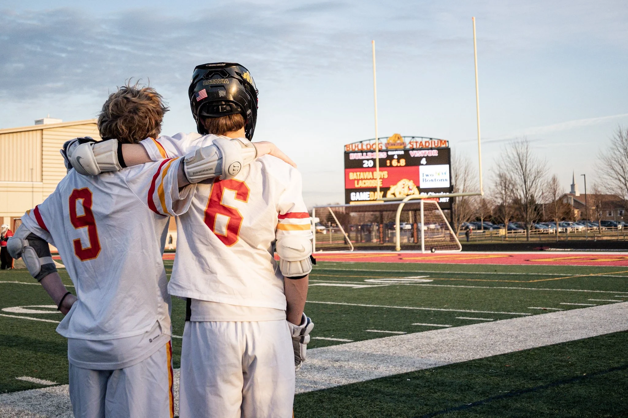 Two football players in white and red uniforms are standing on a football field, with one putting his arm around the other's shoulder, celebrating or comforting. The player on the left is wearing a helmet and has the number 9 on his jersey, while the