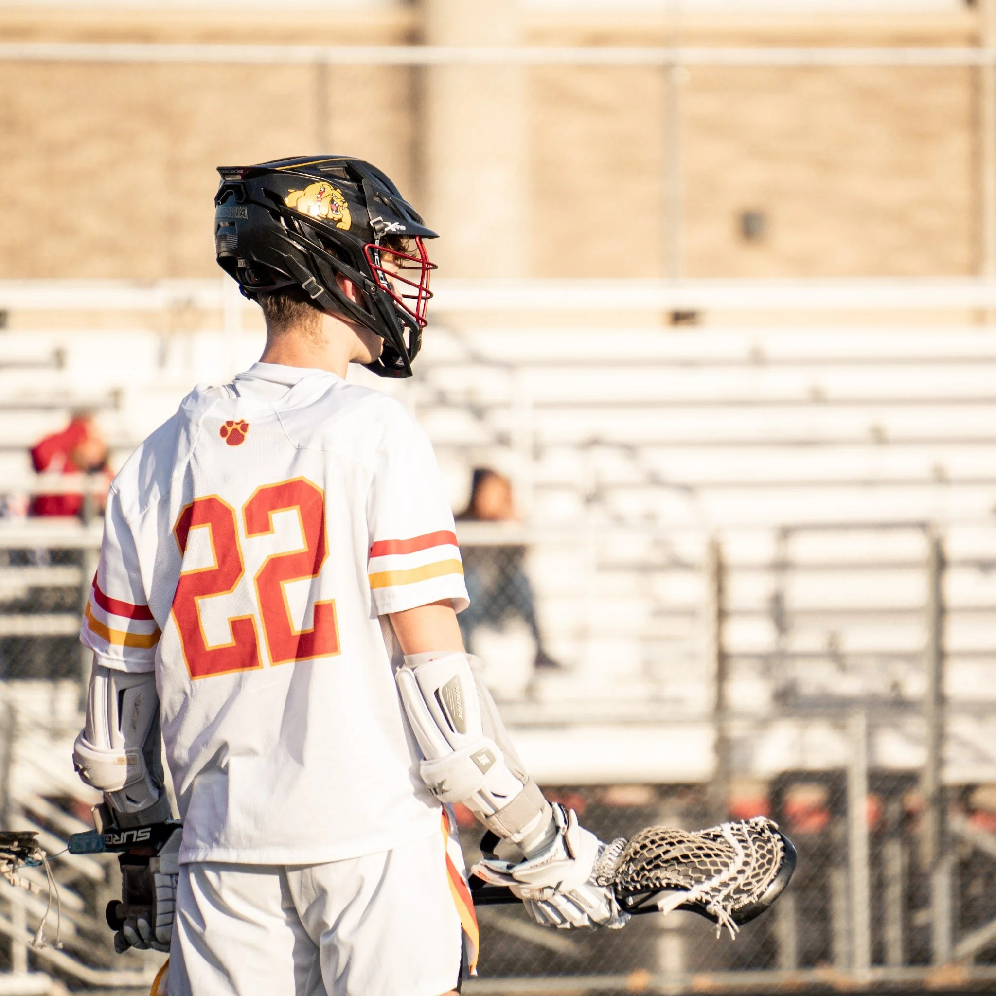 A lacrosse player in a white jersey with red and yellow stripes and the number 22, wearing a black helmet with a red faceguard, holding a lacrosse stick, standing on a sports field.