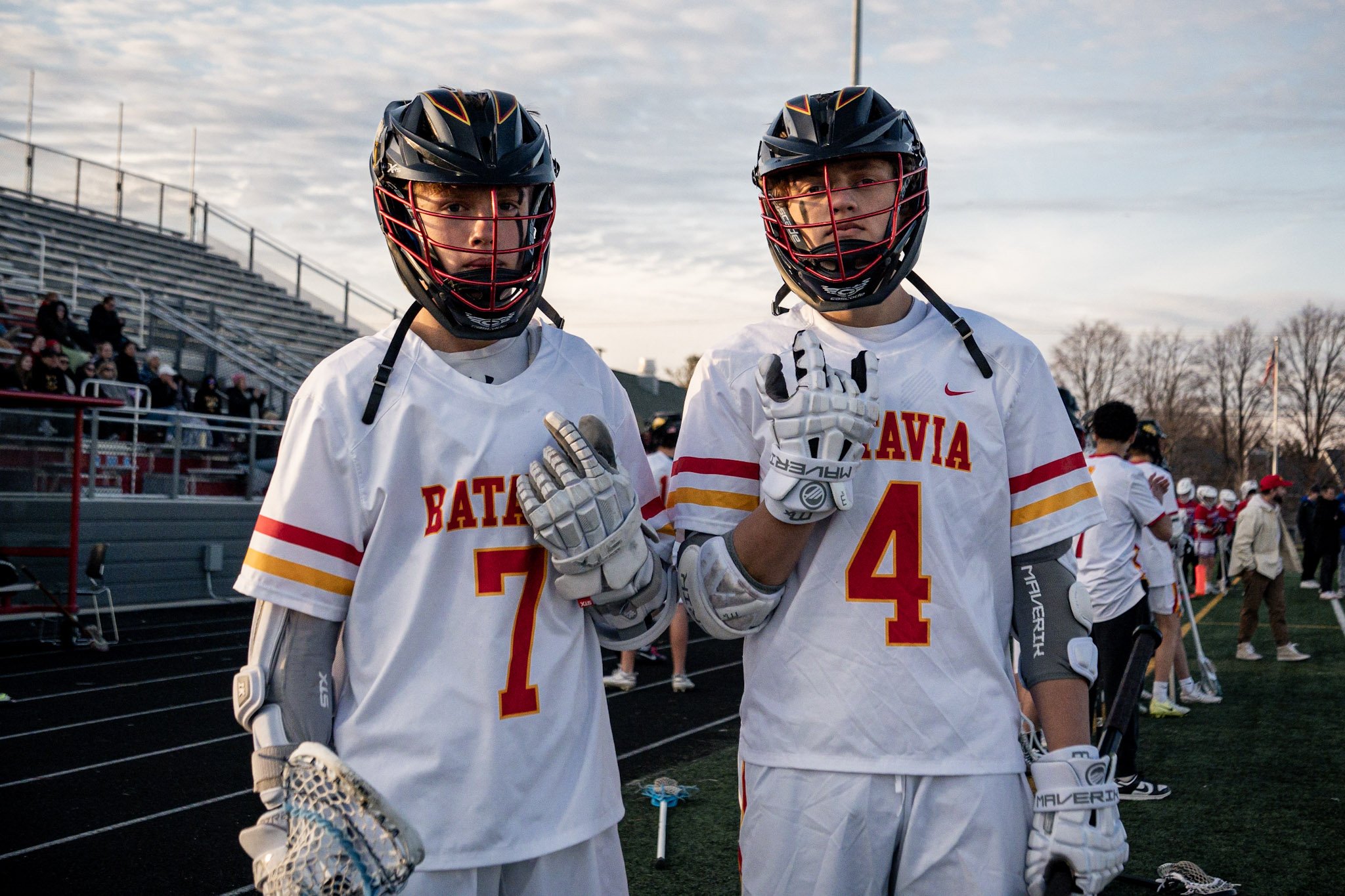 Two male lacrosse players wearing white jerseys with red and yellow accents, helmets with face masks, and gloves on a sports field, giving a peace sign and looking at the camera.