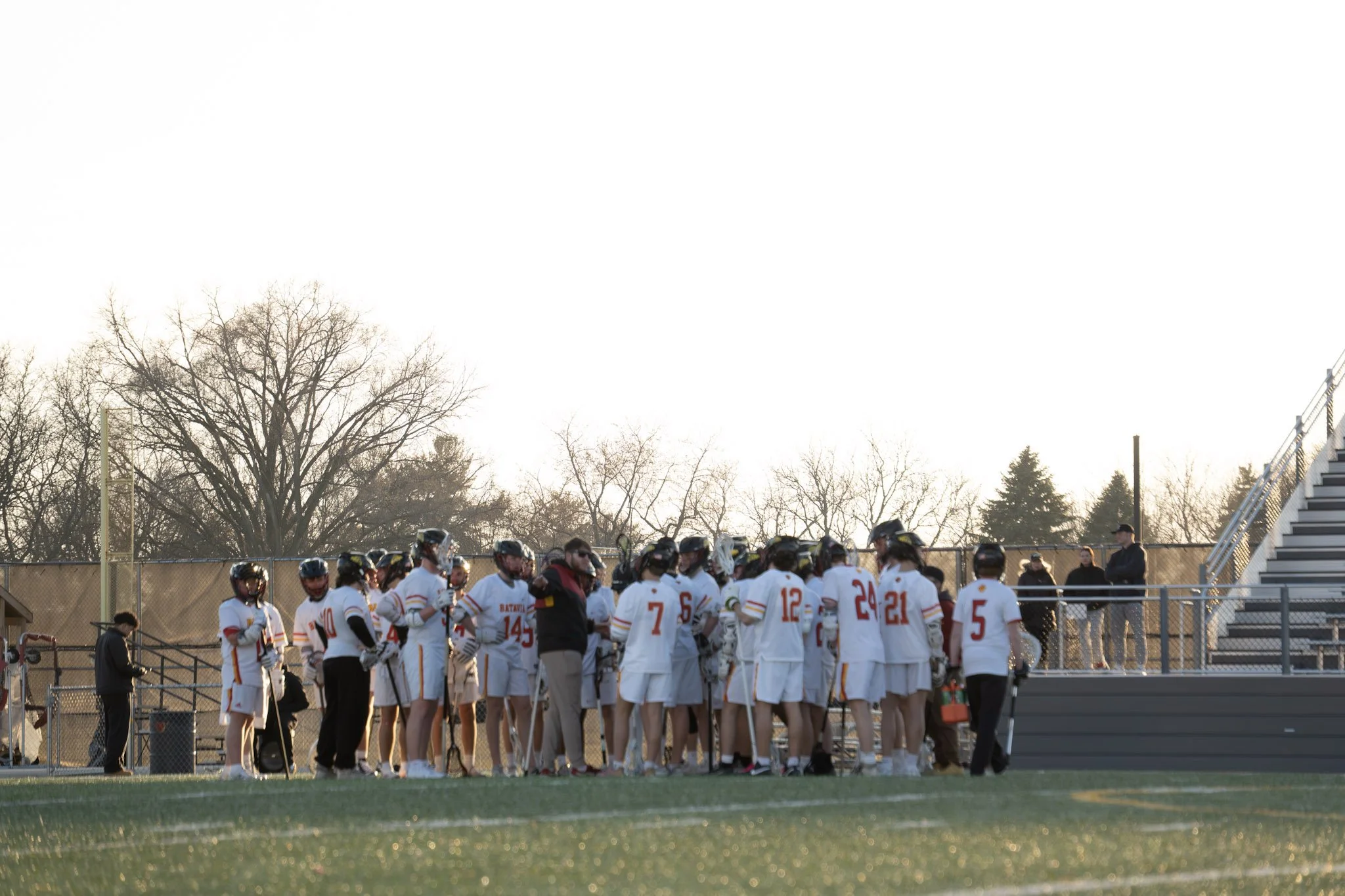 Lacrosse team members in white jerseys gathered in a huddle on the field, listening to their coach during a game or practice, with some people standing along the sidelines and empty bleachers in the background.