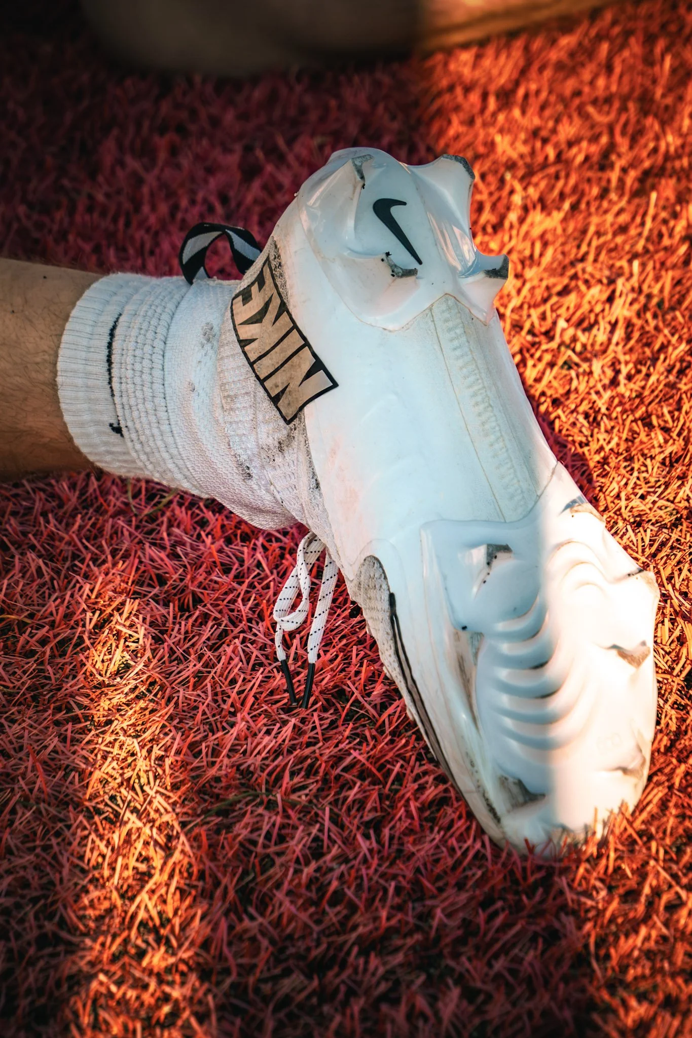 A white Nike football cleat resting on a red turf field, with a white sock beneath it.