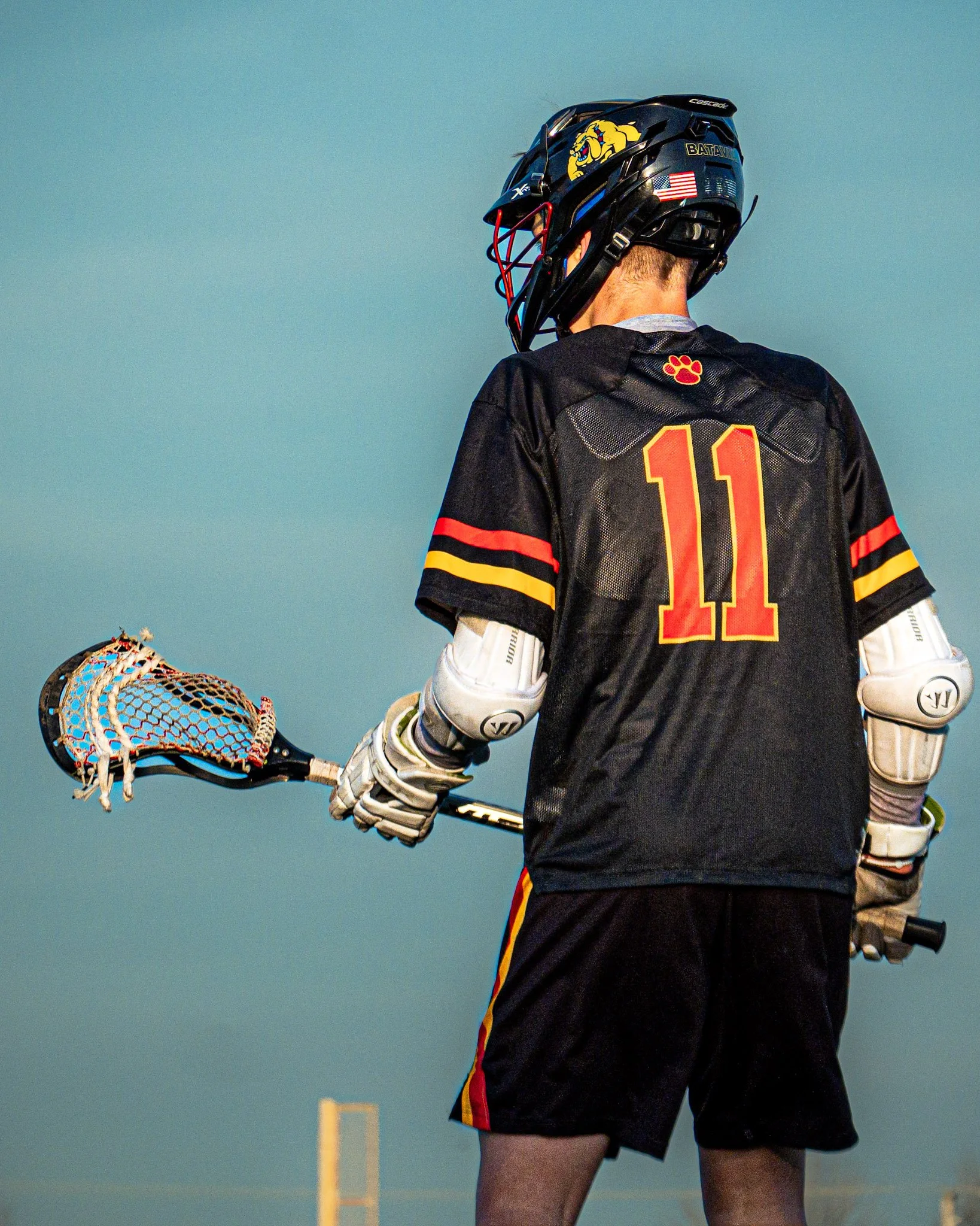 A lacrosse player dressed in a black jersey with the number 11, wearing a lacrosse helmet and gloves, holding a lacrosse stick, standing outdoors against a blue sky background.