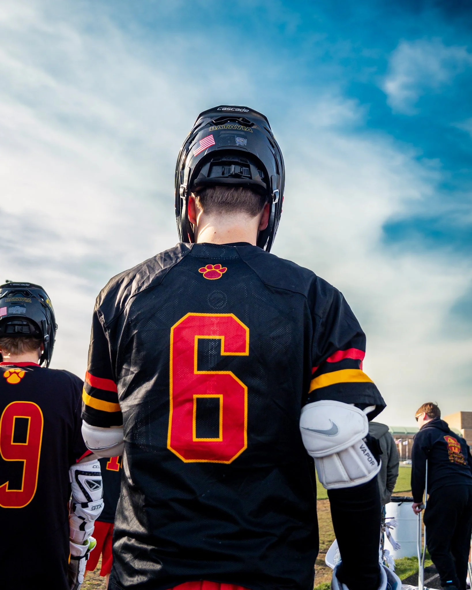 Back view of an American football player wearing a black jersey with number 6, red and yellow accents, and a helmet with an American flag decal, standing on a field with other players and a partly cloudy sky.