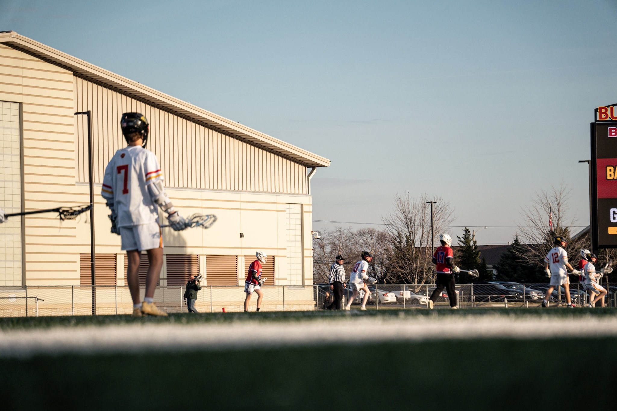 Lacrosse players on the field during a game, with a large building and scoreboard in the background.