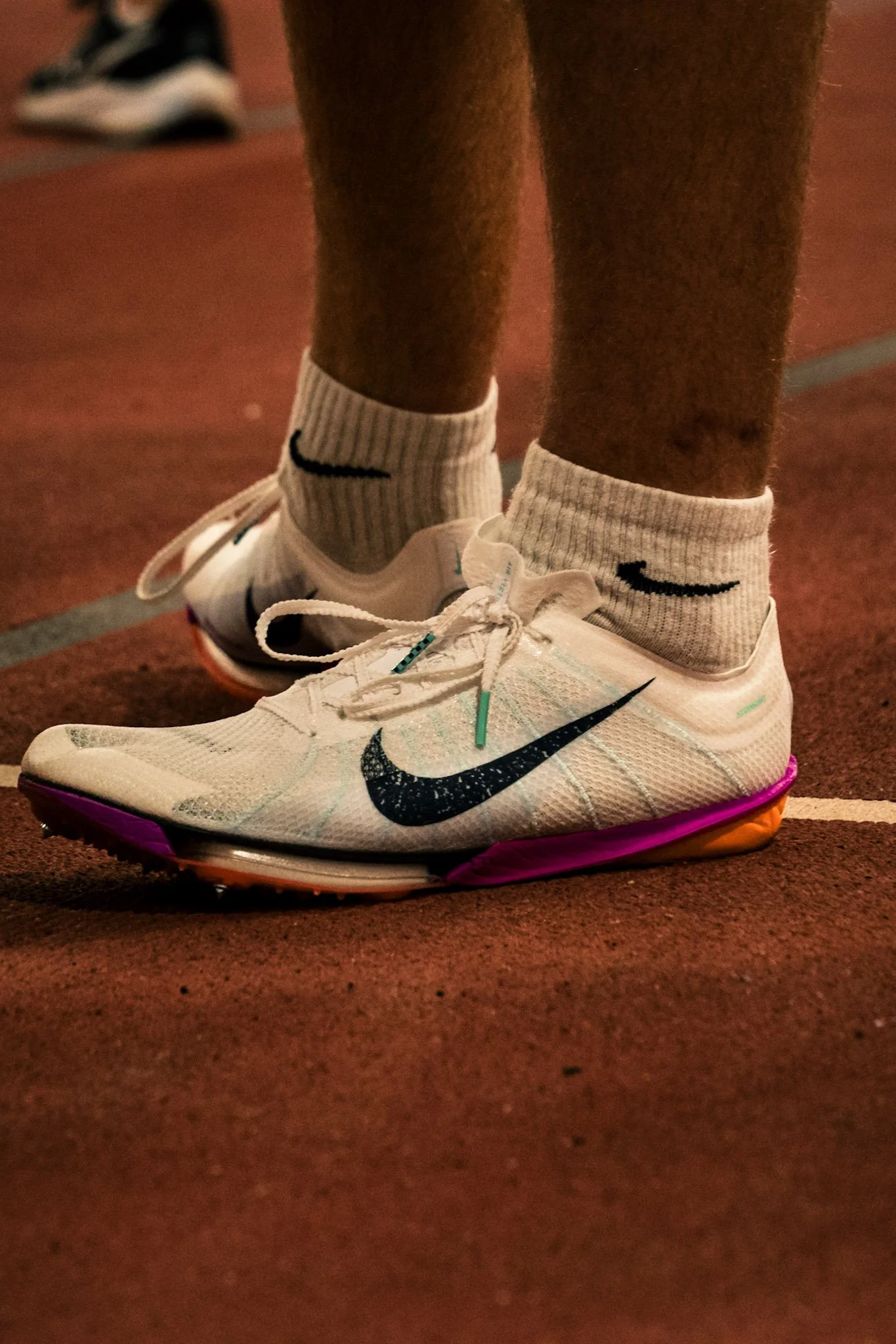 Close-up of a person wearing white Nike athletic shoes with black swoosh logos, white Nike socks with black swoosh, and standing on a reddish running track.