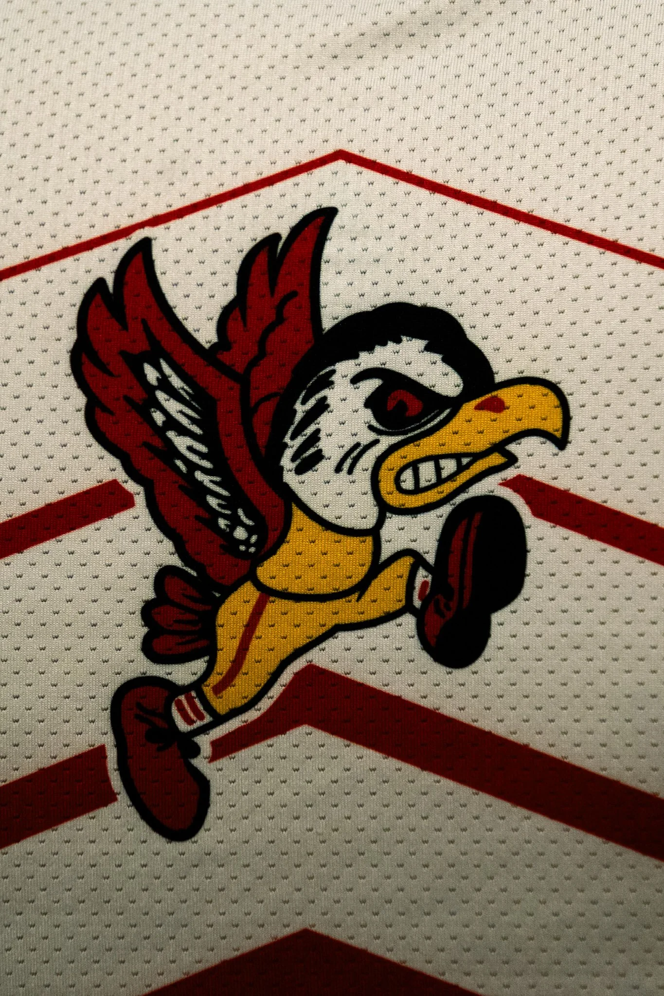 A cartoon mascot of a duck with red wings, a yellow body, and a black and white head, running with a determined expression on a sports jersey fabric with red stripes.