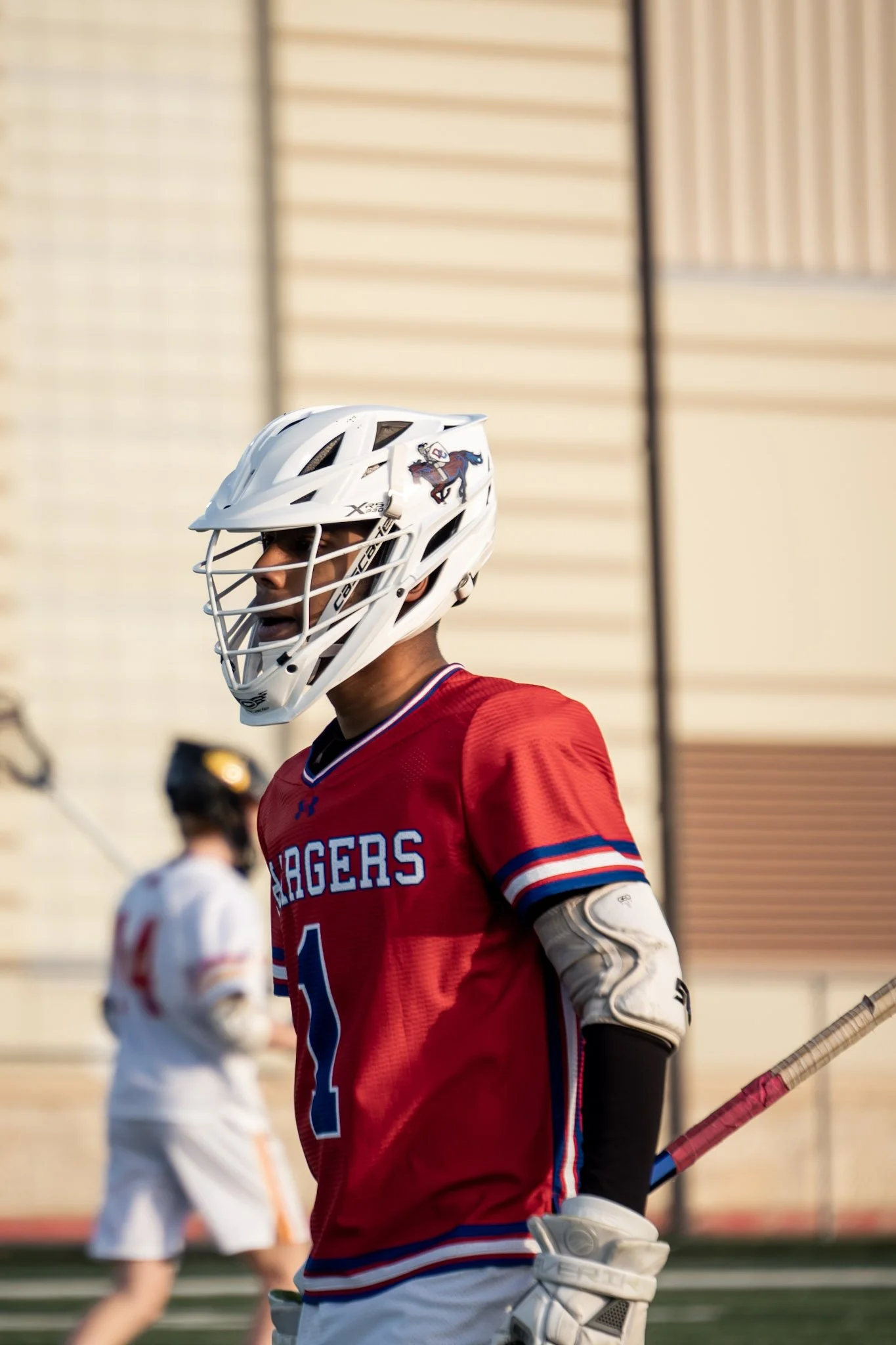 Lacrosse player wearing a red jersey with number 1, white helmet with a horse logo, and holding a lacrosse stick on a field, with another player in the background.