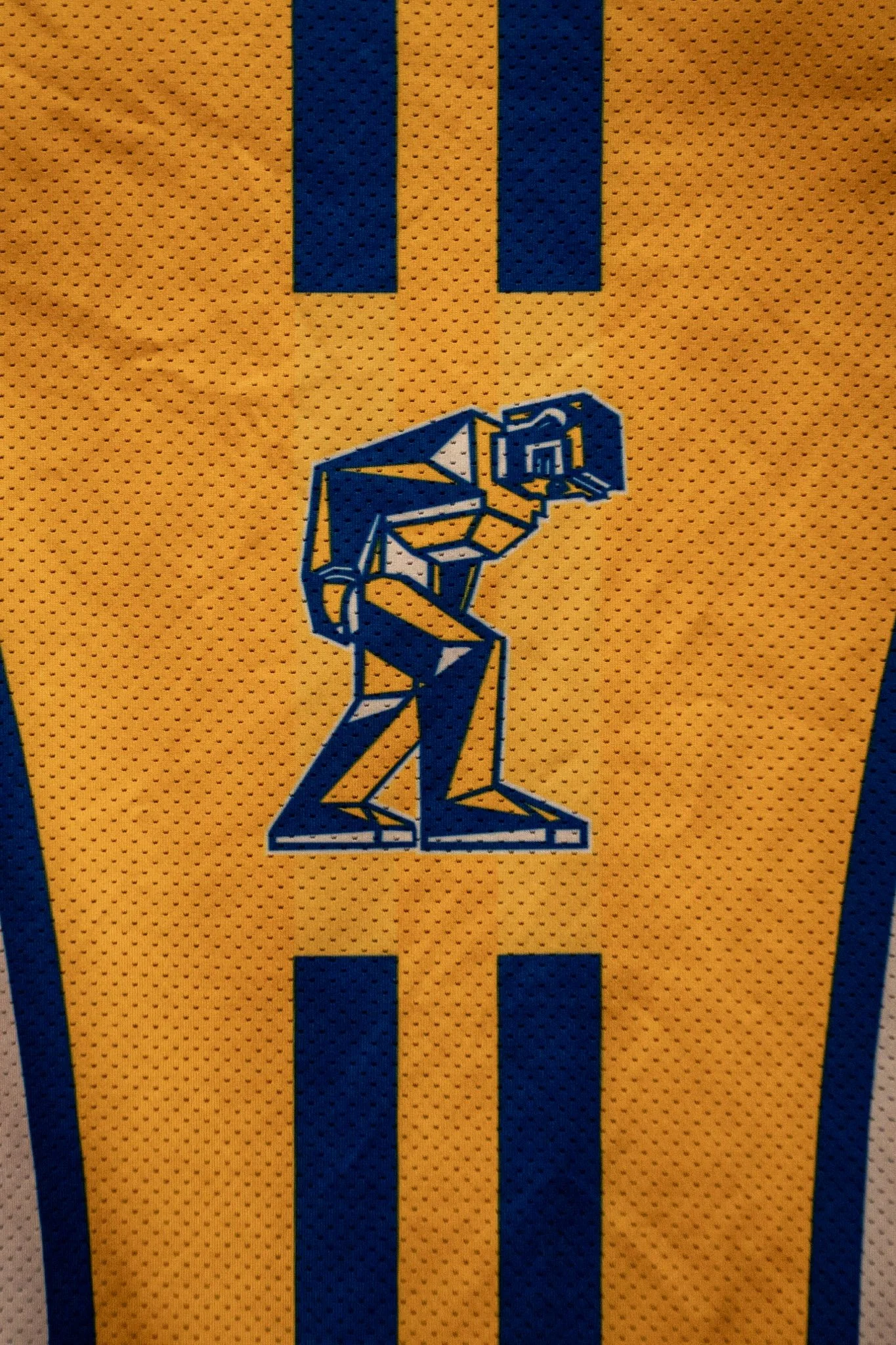 Close-up of a yellow sports jersey with a blue geometric robot logo and blue stripes.
