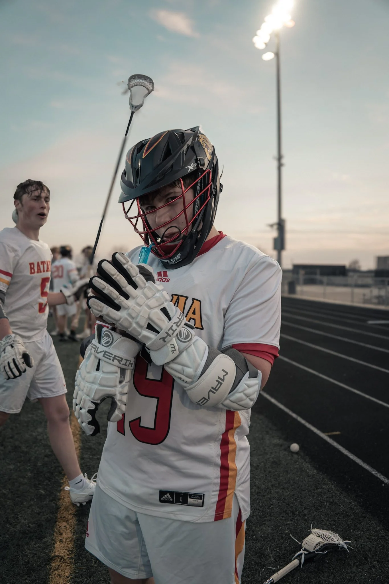 Lacrosse player wearing a helmet and gloves, holding his lacrosse stick with a is open-mouthed, excited expression on his face during a game at sunset on the field.