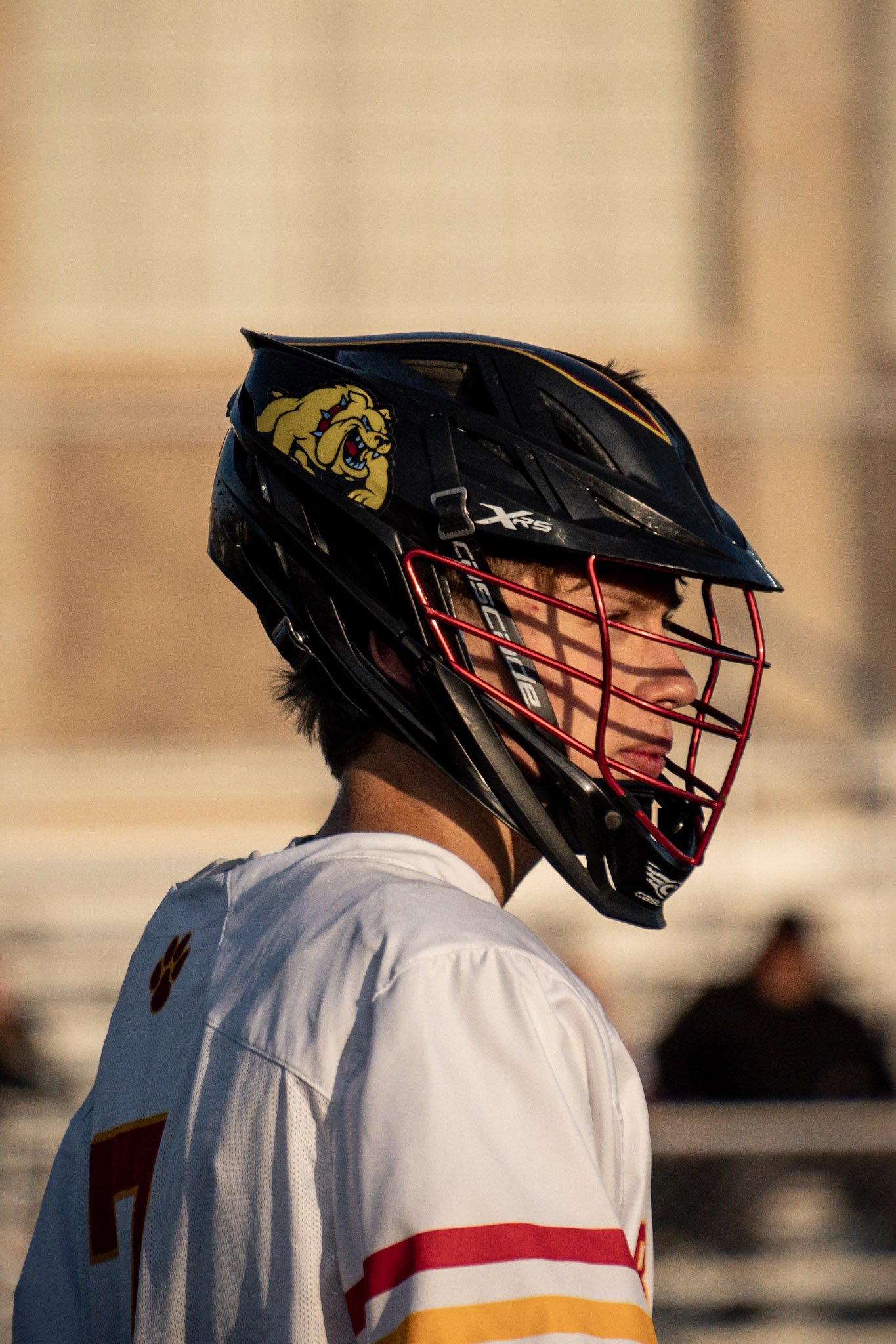 A male athlete wearing a black lacrosse helmet with a yellow lion logo and a white jersey with red and yellow stripes.