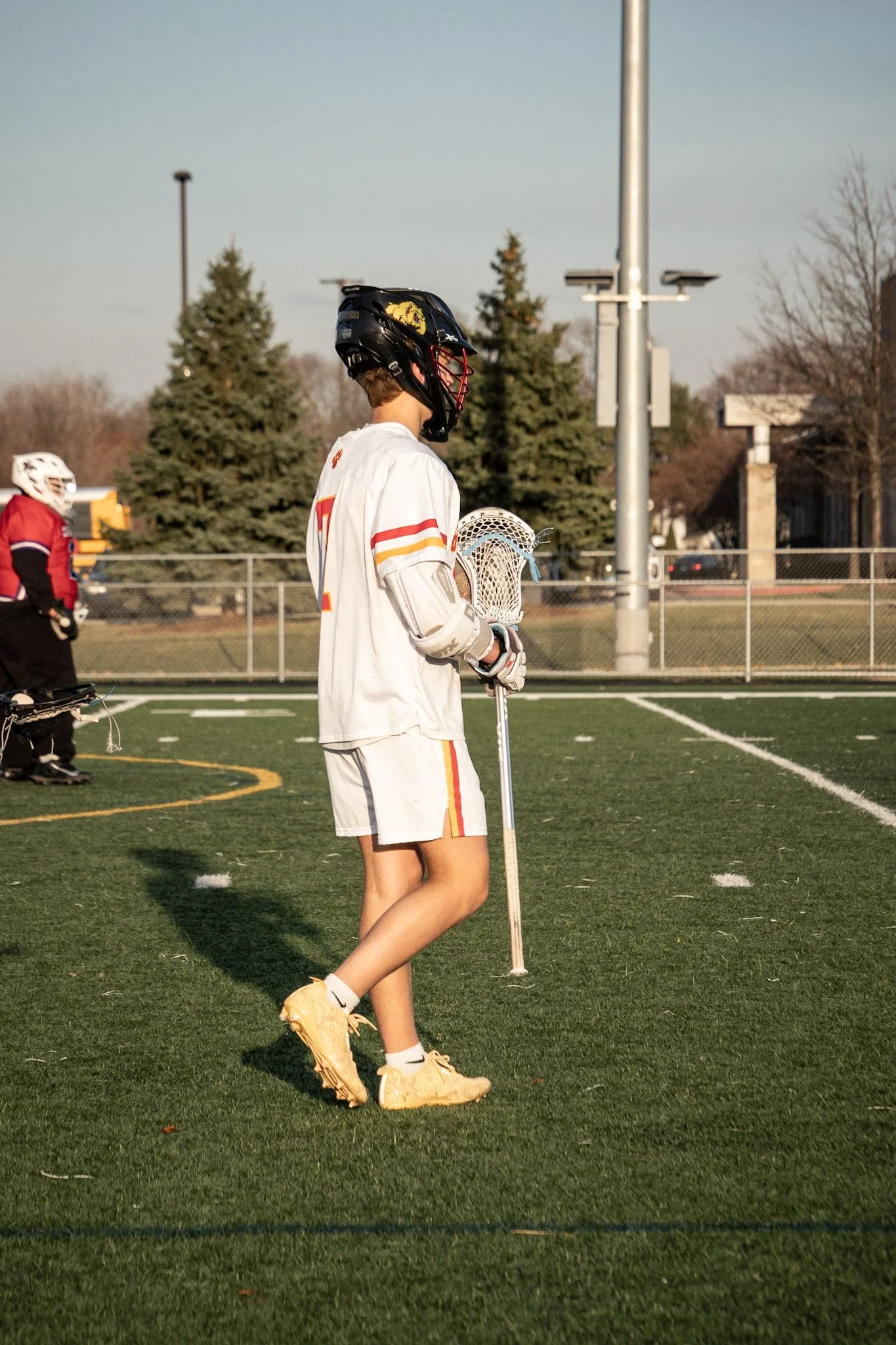 Lacrosse player standing on a field holding a lacrosse stick, wearing a helmet, white uniform with red and yellow stripes, yellow shoes, and facing away from the camera.