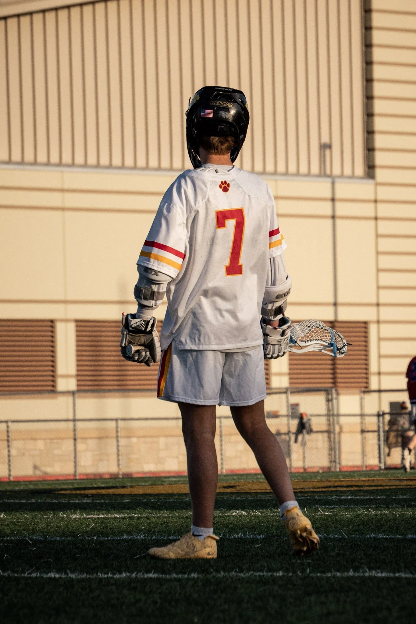 Lacrosse player in white uniform with red and yellow accents, number 7, standing on the field with helmet and gloves, holding a lacrosse stick, during sunset.