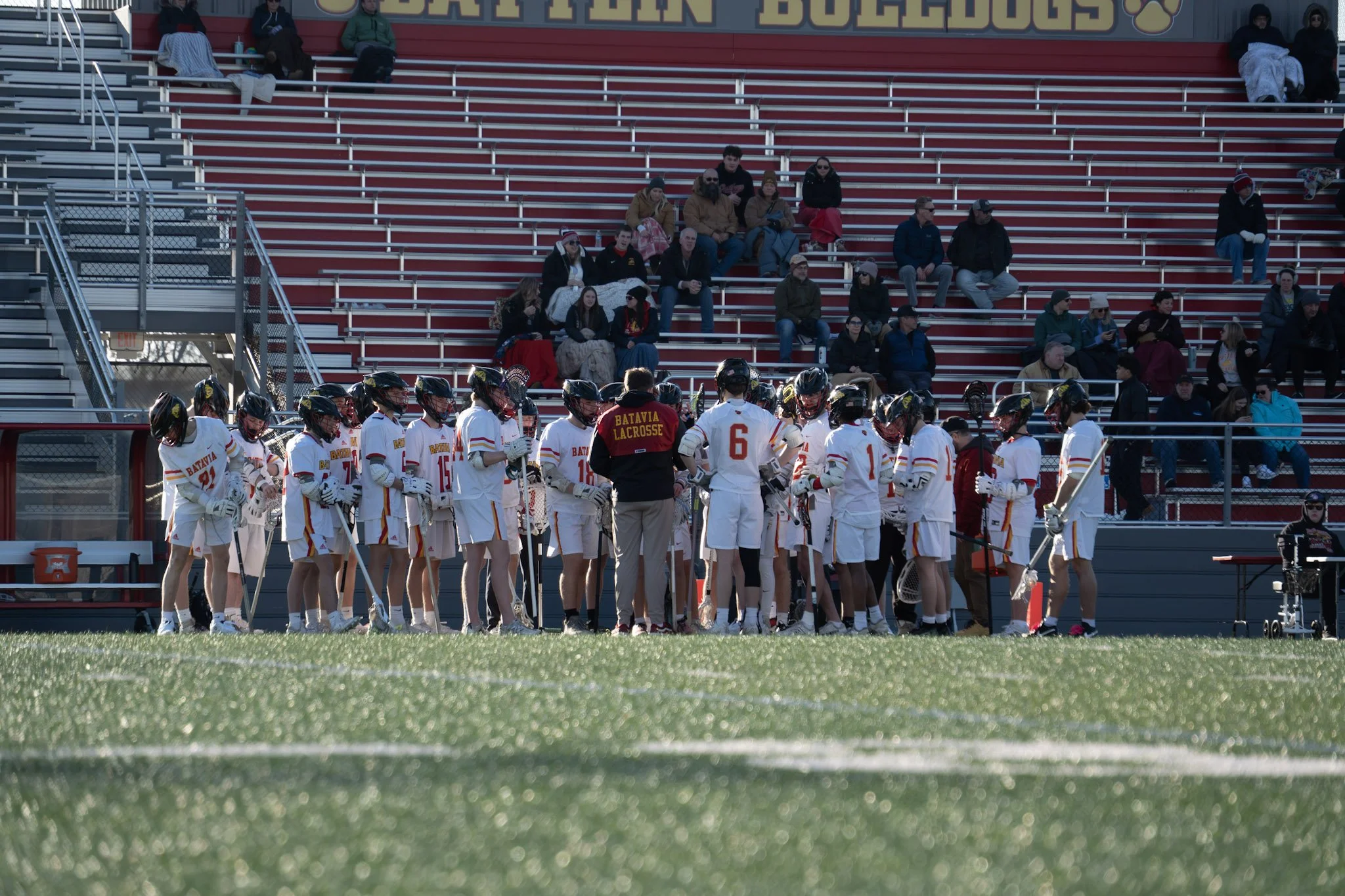 Lacrosse team dressed in white uniforms with red and gold accents, huddled on the field, with spectators sitting on bleachers in the background at a stadium.