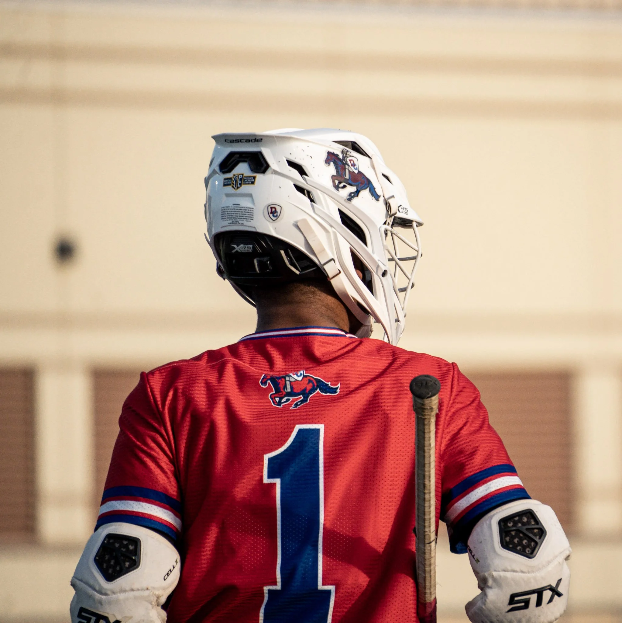 Hockey player wearing a red jersey with a blue number 1 and a horse logo, holding a hockey stick, viewed from behind, with a white helmet.