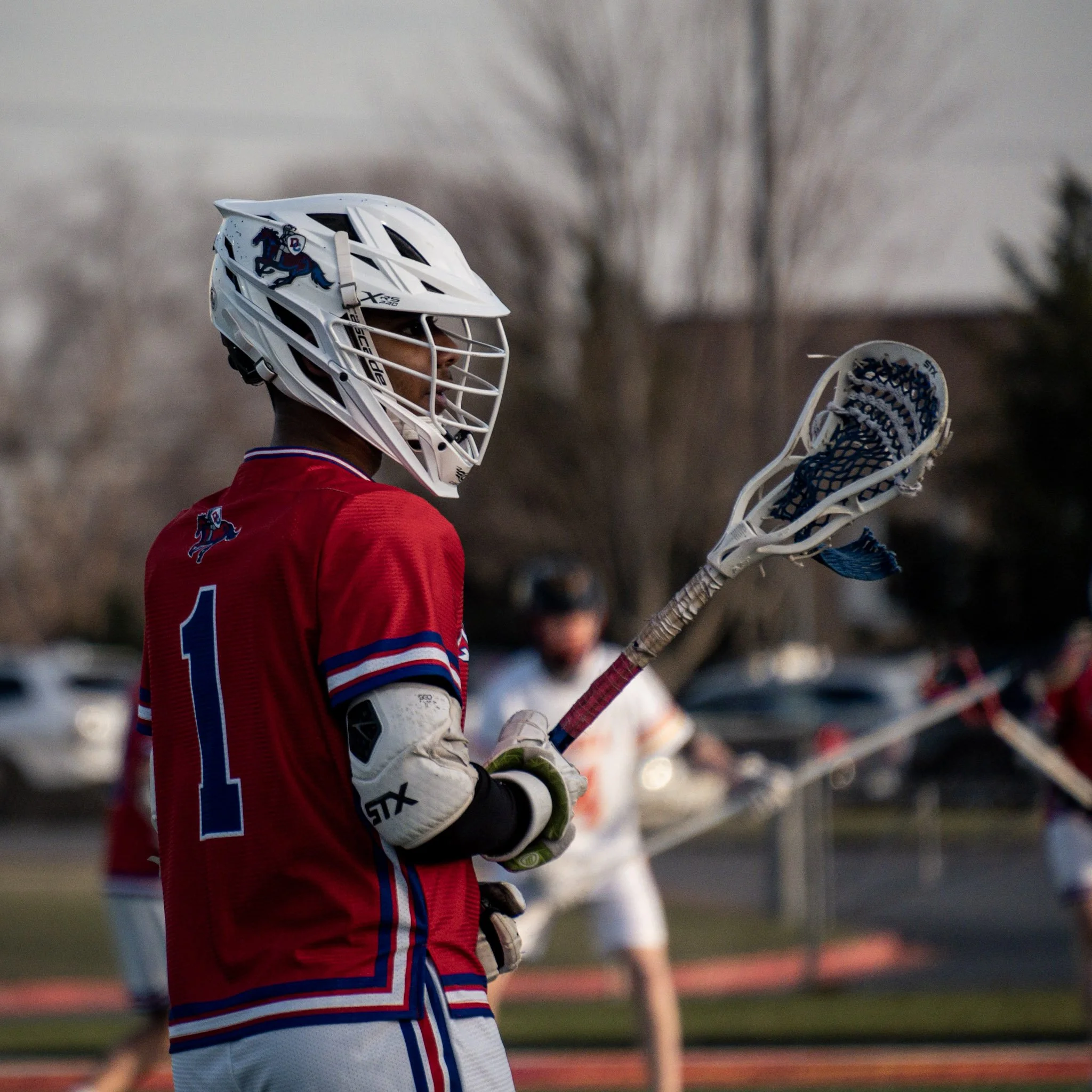 Lacrosse player wearing a red jersey and white helmet holding a lacrosse stick during a game.