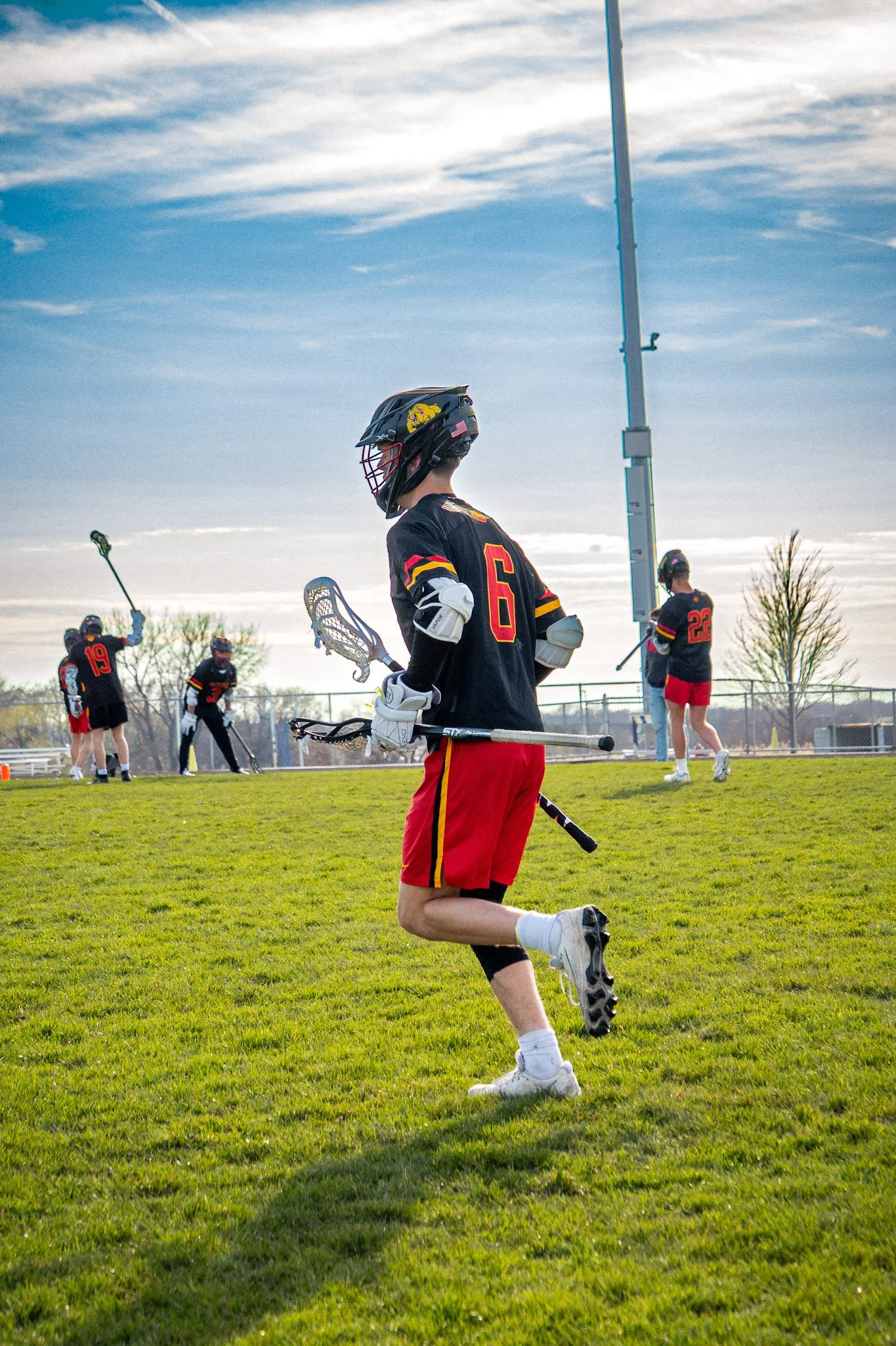 Lacrosse players on a grassy field wearing black jerseys with red and yellow accents, with a blue sky and trees in the background.