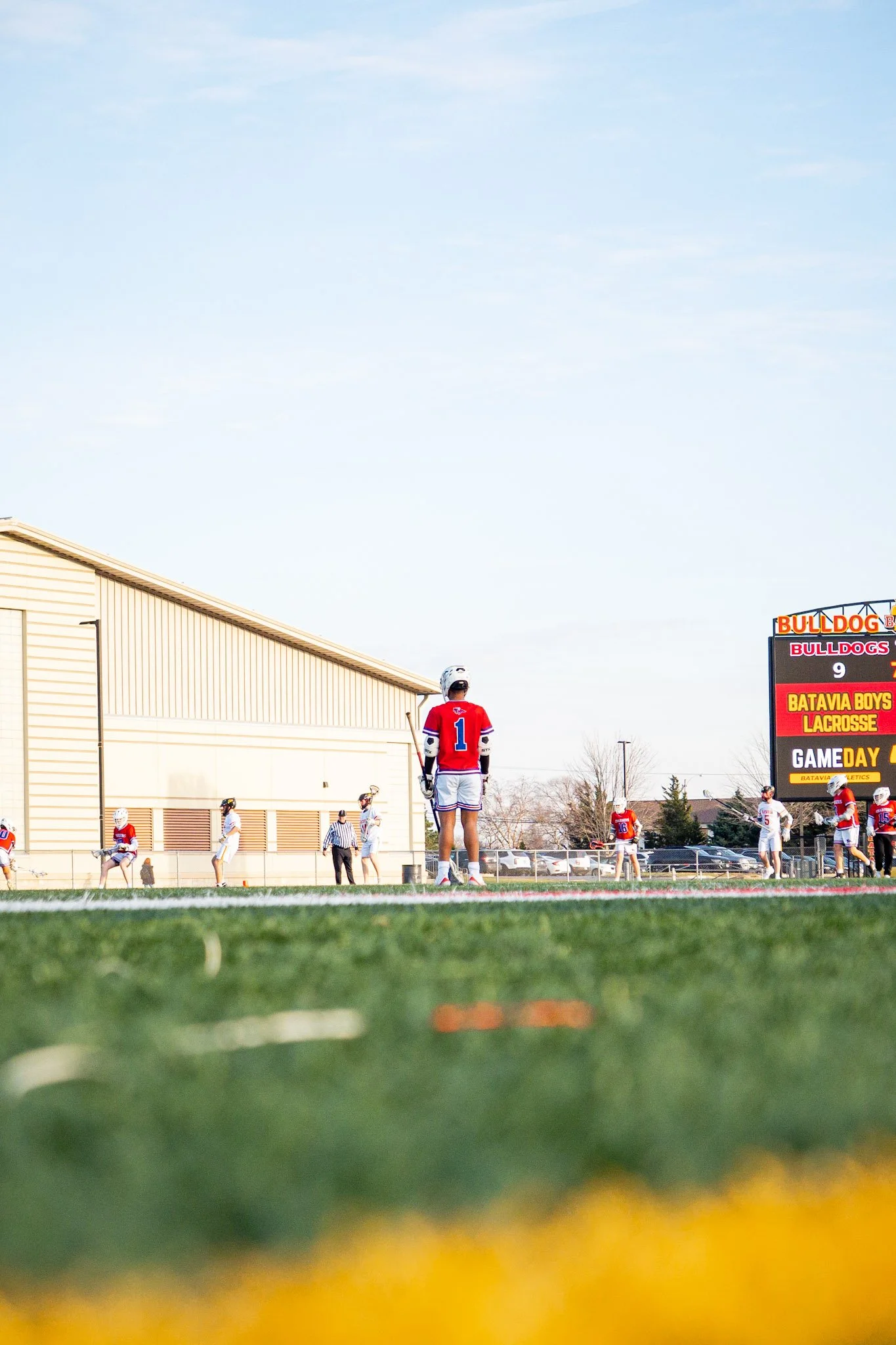 A lacrosse game in progress on a field near a beige building with players in red and white uniforms, some standing and others preparing to play, with a digital scoreboard showing team names and scores.