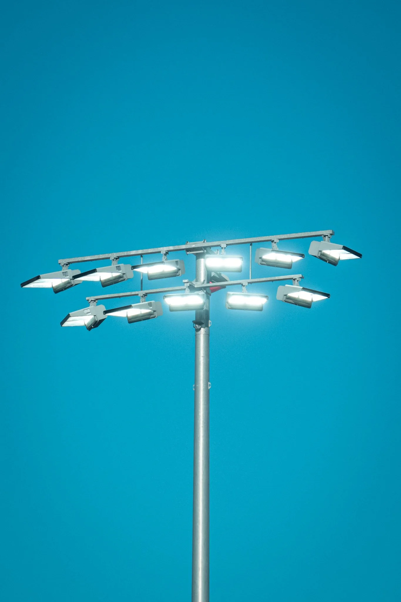 A tall stadium floodlight pole with multiple lights turned on under a clear blue sky.