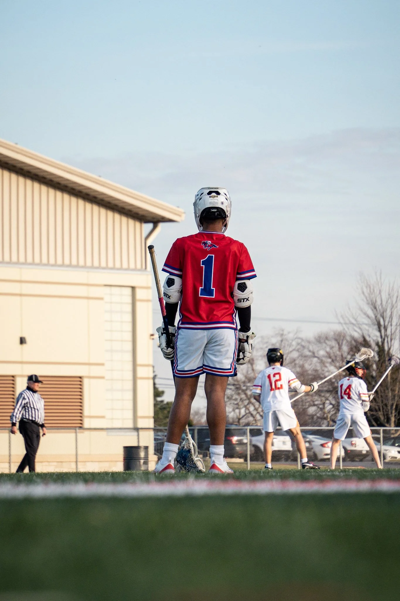 Lacrosse players on the field, with one player in a red jersey numbered 1 prominently in the foreground, holding a lacrosse stick, and other players and a referee in the background.