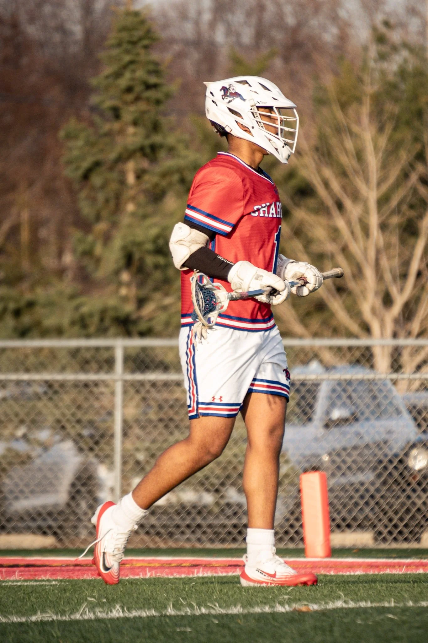 A lacrosse player wearing a red jersey, white shorts with matching red and blue stripe details, white cleats, and gloves, holding a lacrosse stick, and wearing a white helmet, walking on the field during a game.