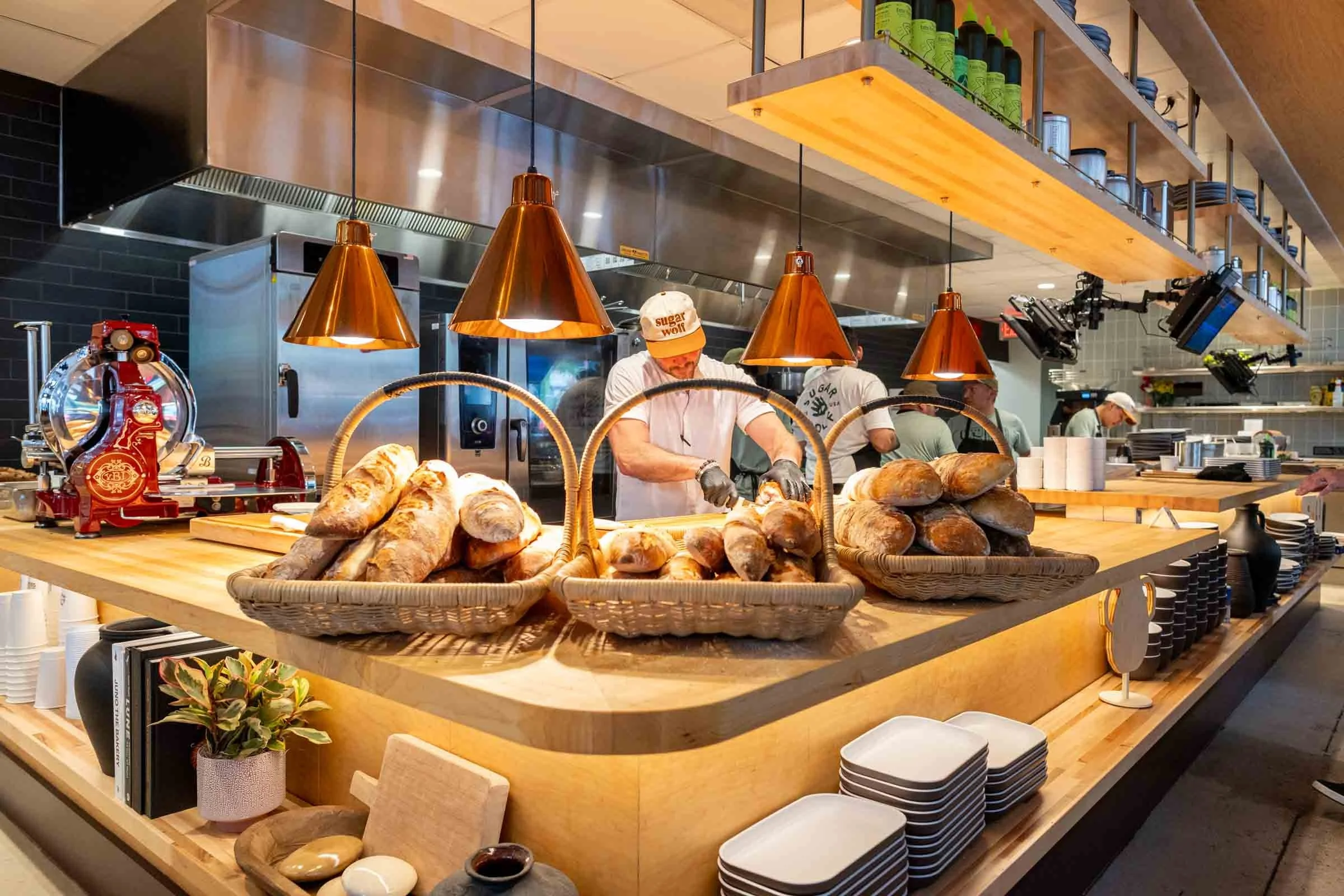 A modern restaurant kitchen with a wooden serving counter filled with bread in baskets. There are copper pendant lights hanging from the ceiling, chefs working behind the counter, and a shelf with dishes and supplies above.