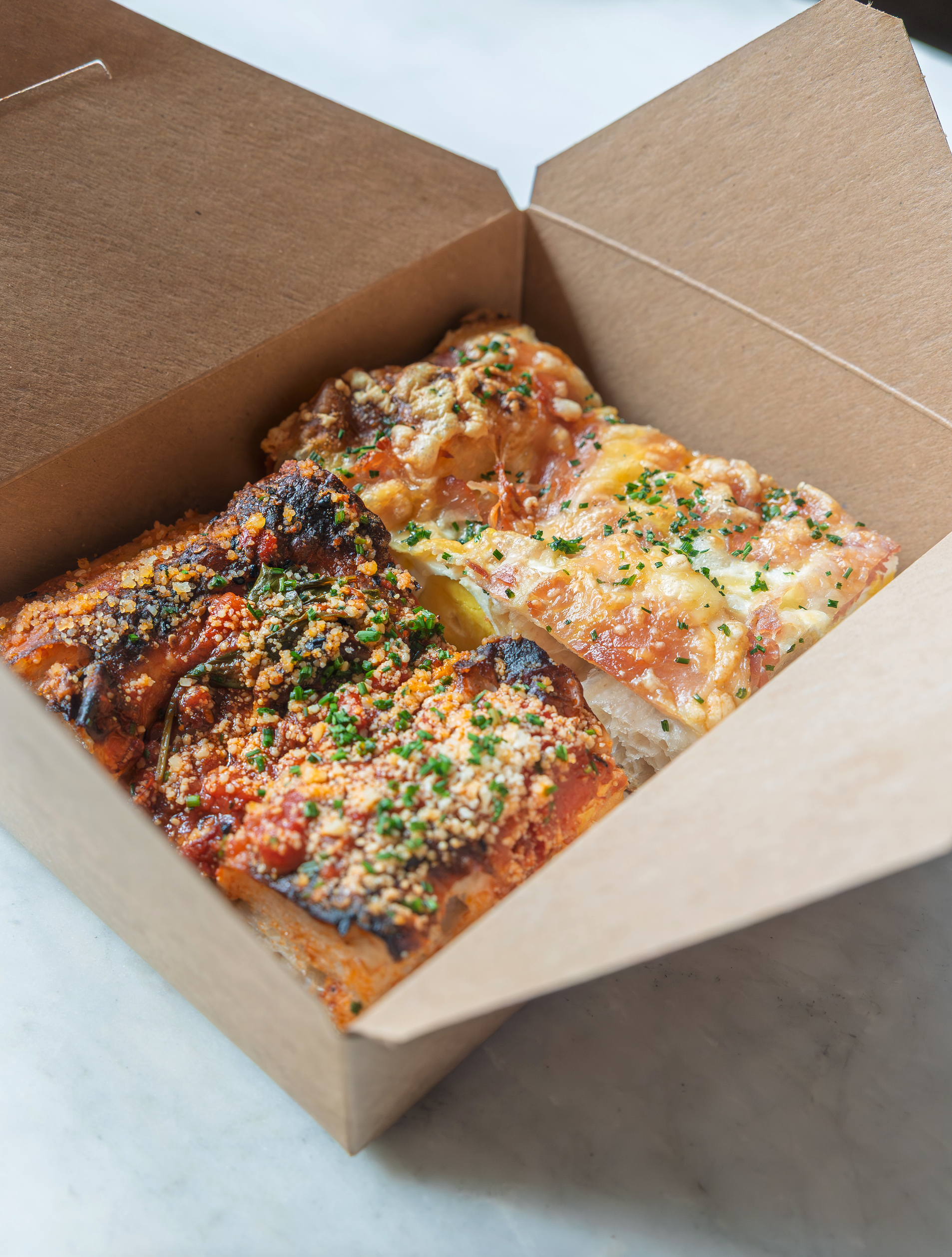 Two pieces of lasagna in a brown takeout box, garnished with chopped herbs.