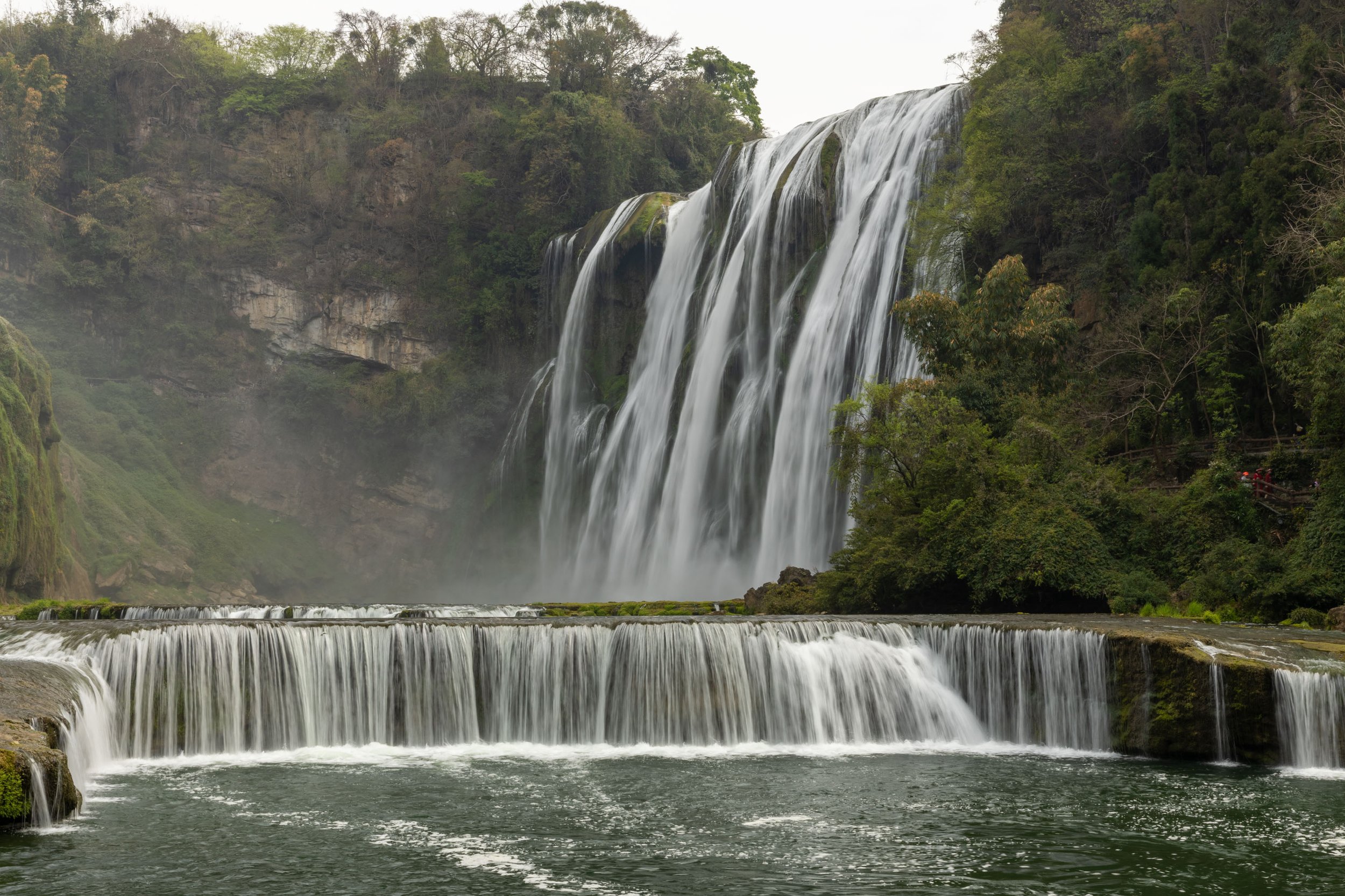 Waterfall cascading over rocks surrounded by lush greenery and forested cliffs.