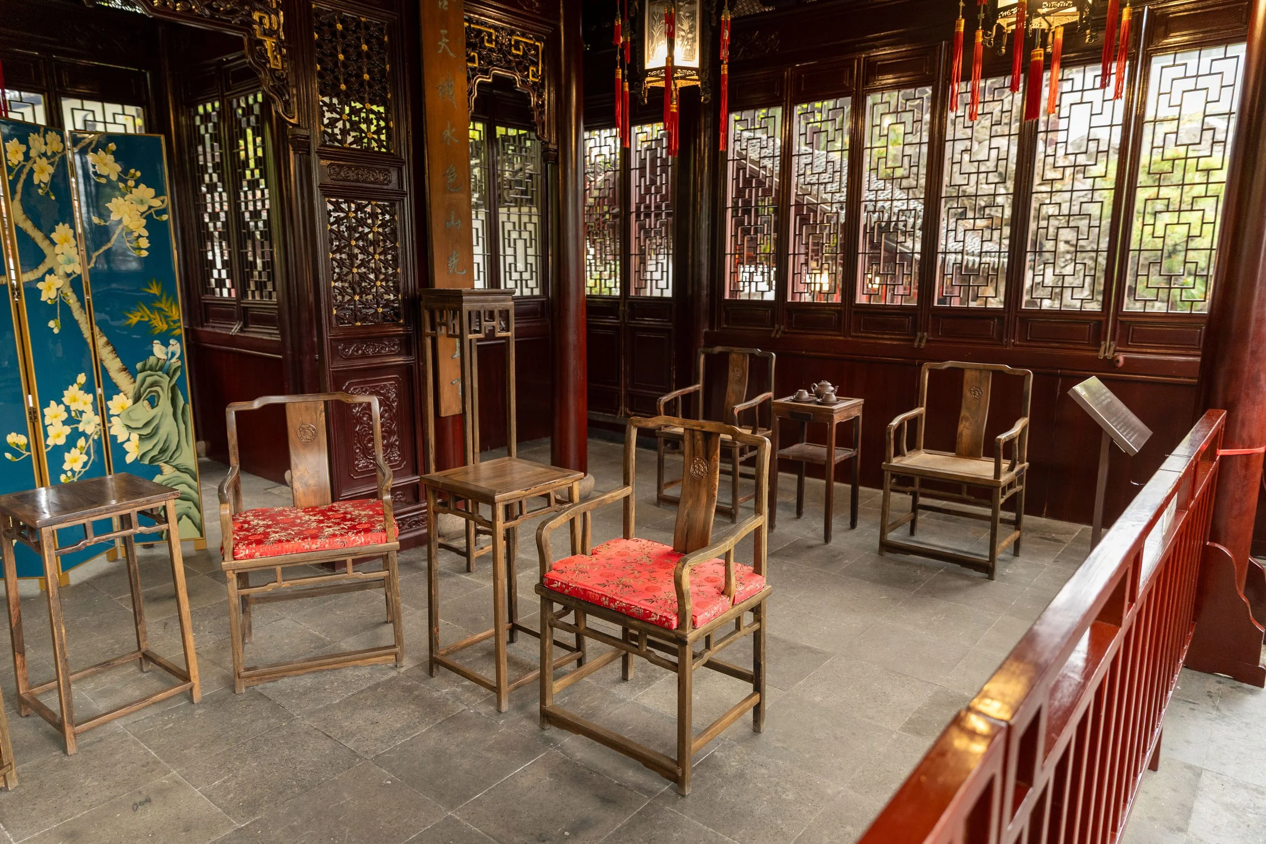 Traditional Chinese room with wooden chairs, stools, and lattice windows; red cushions on chairs; decorative screen and teapot on table.