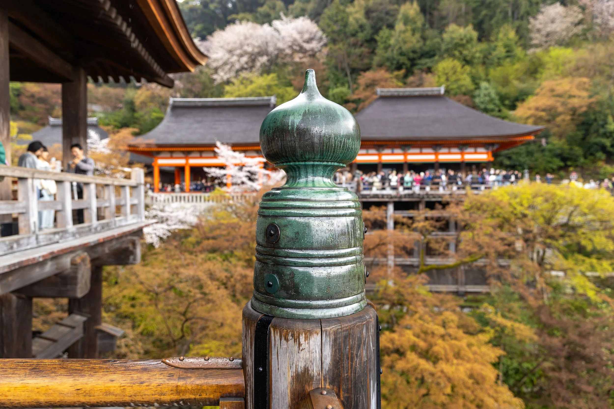 Close-up of a decorative post on a railing in a traditional Japanese temple setting, with blurred background showing temple structures and foliage.