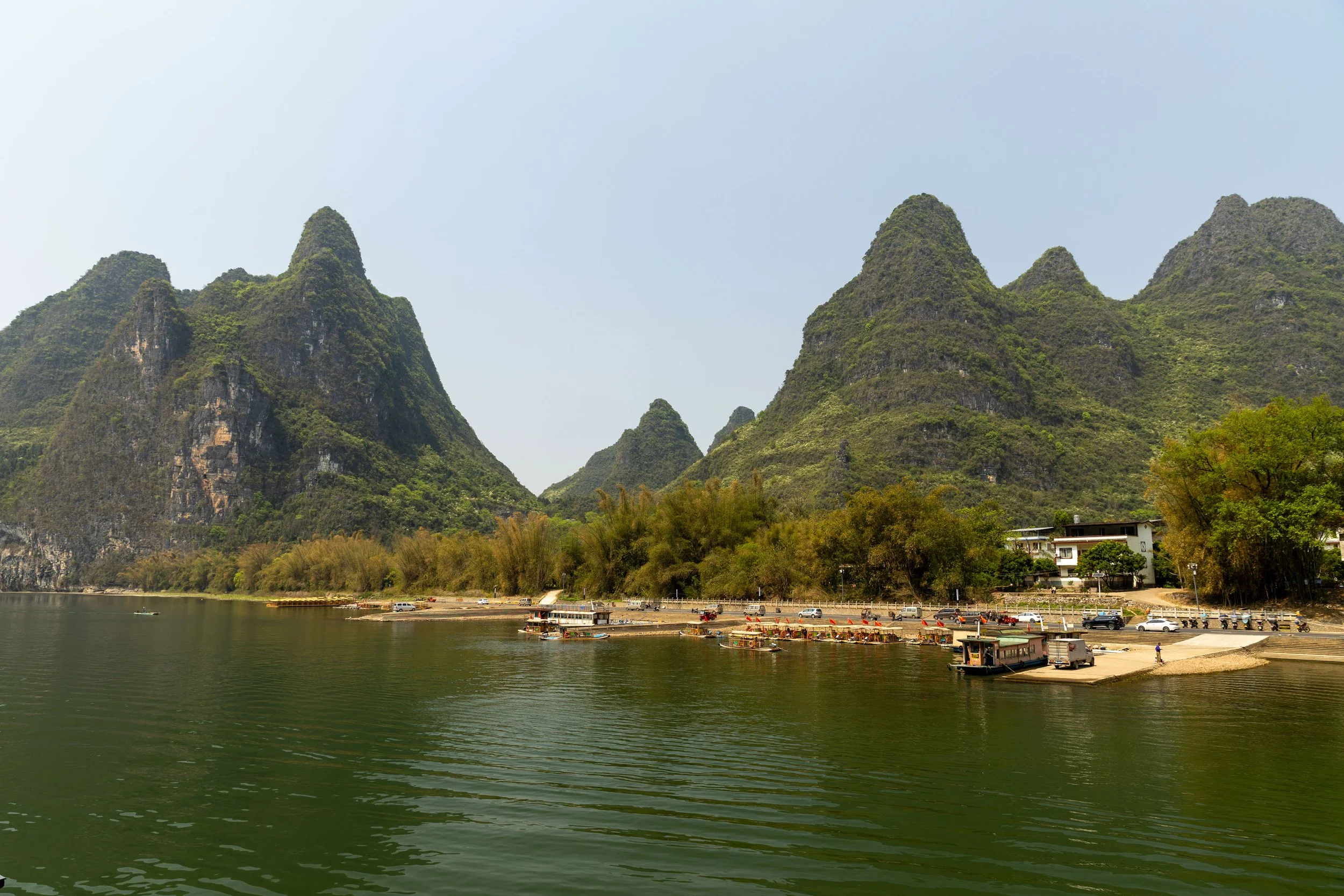 Scenic view of lush karst mountains by a river with boats on the water and a small dock area surrounded by trees.