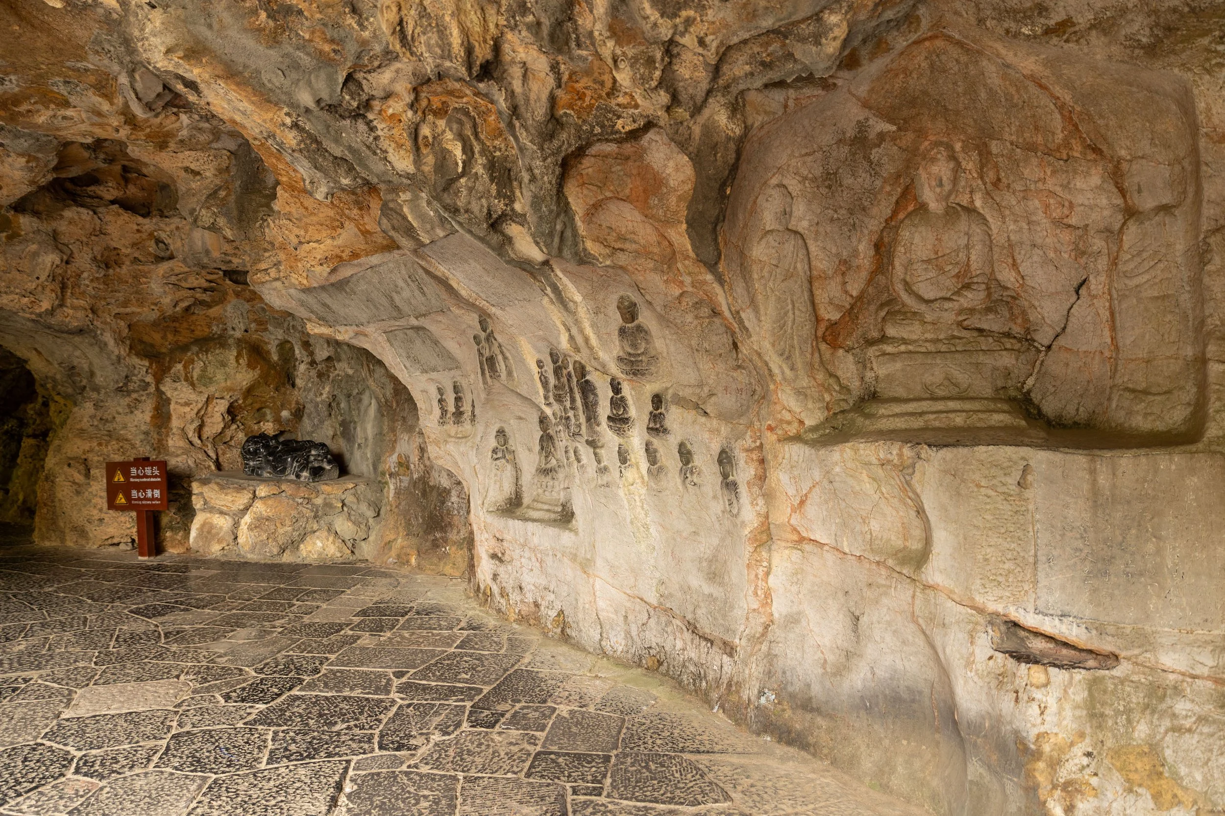 Ancient cave with Buddhist carvings on stone walls, featuring multiple Buddha figures and a small sculpture. Sign in Asian script is visible.