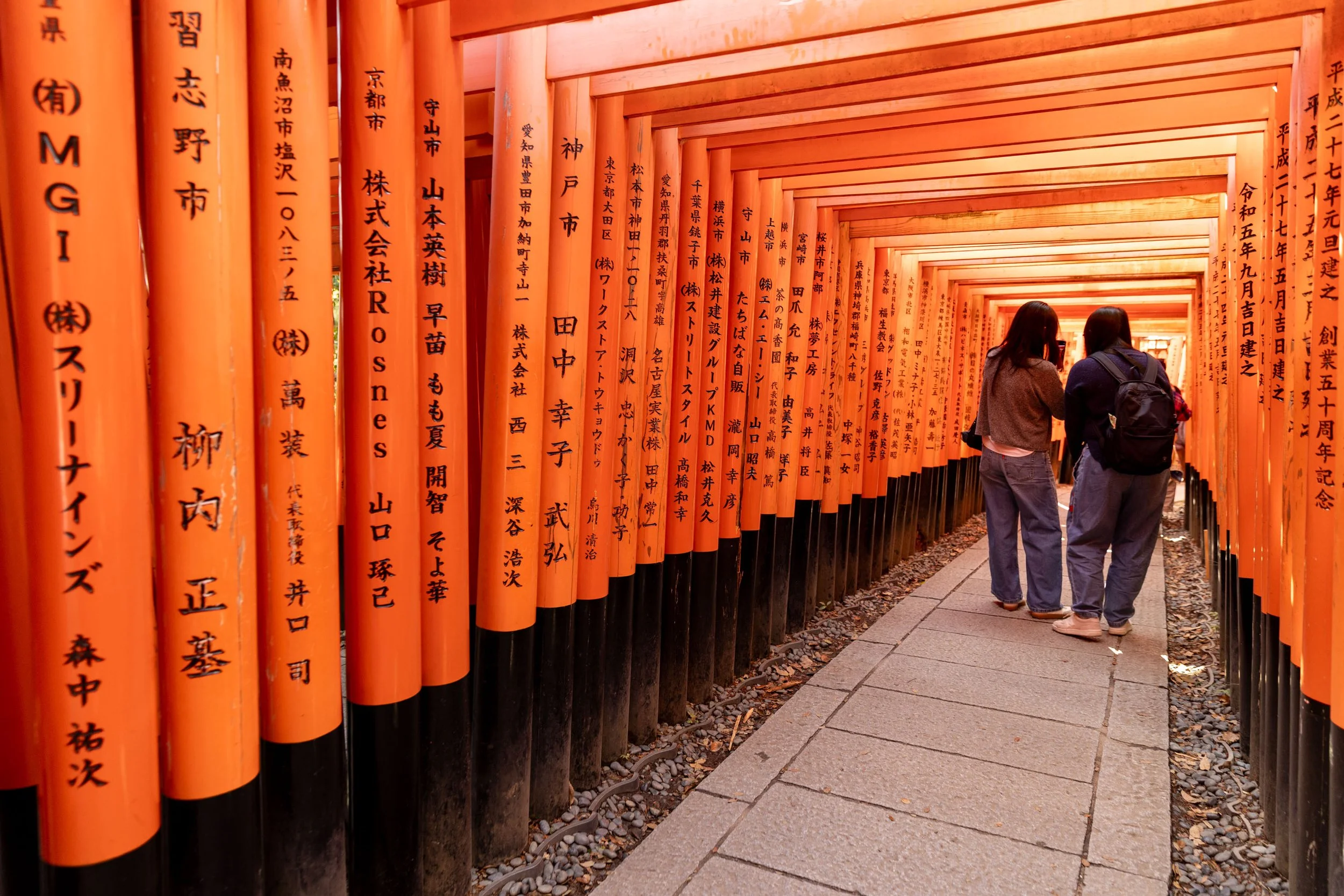 Orange torii gates with black inscriptions at Fushimi Inari Shrine, Kyoto, with two people walking through.
