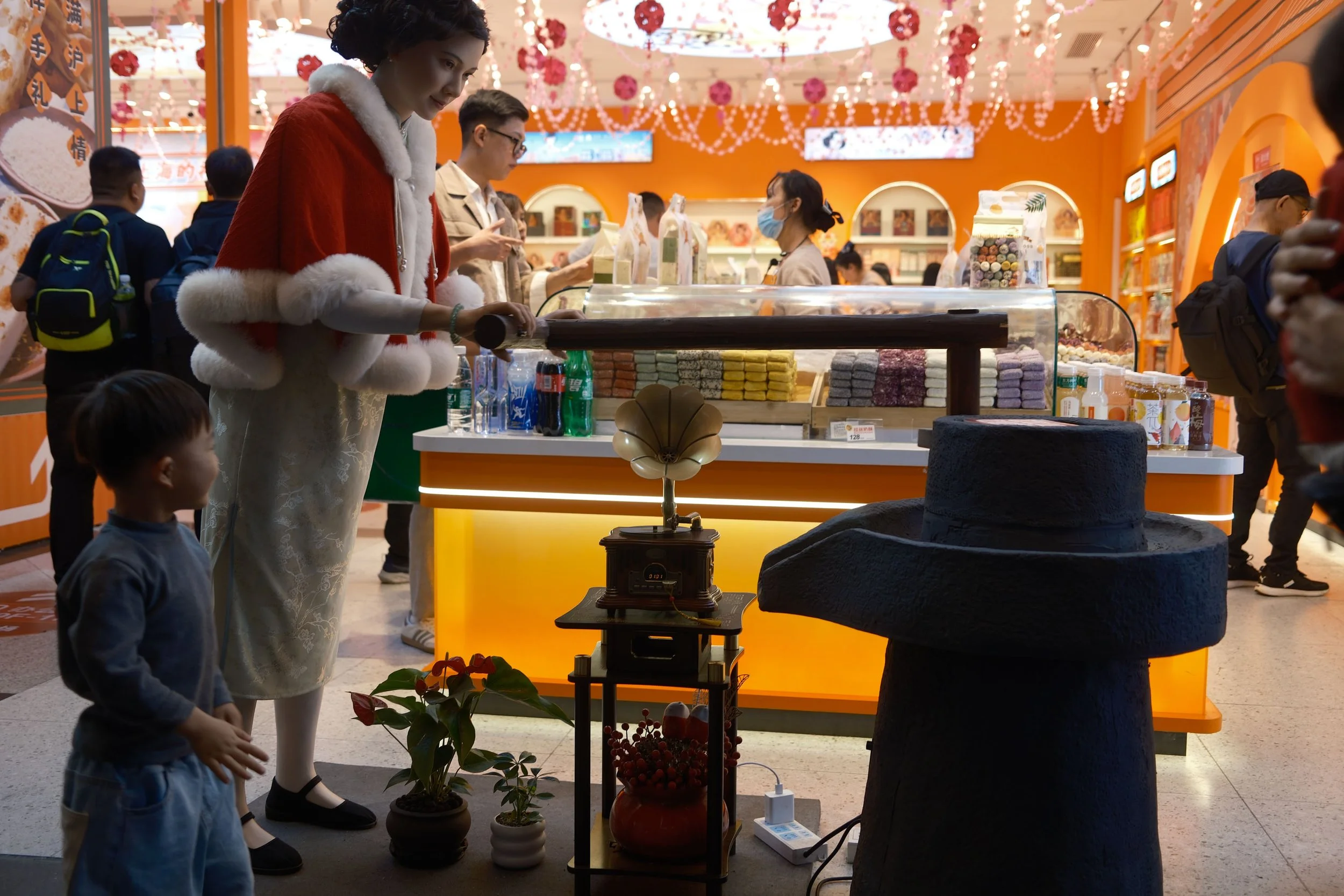 Store interior with mannequin in red cape, table with ceramic pots and gramophone, and people shopping in background.