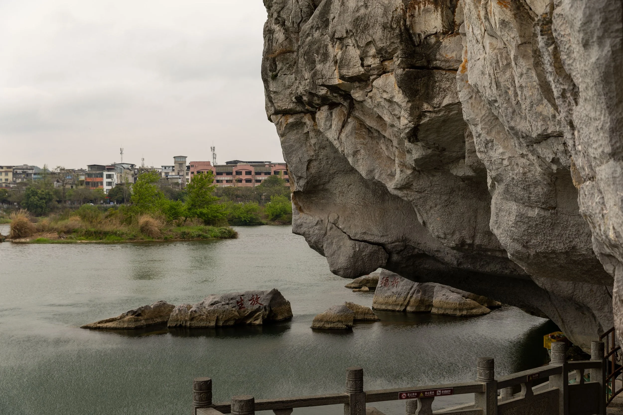 Cave overlooking a river with large rocks and distant buildings in the background.