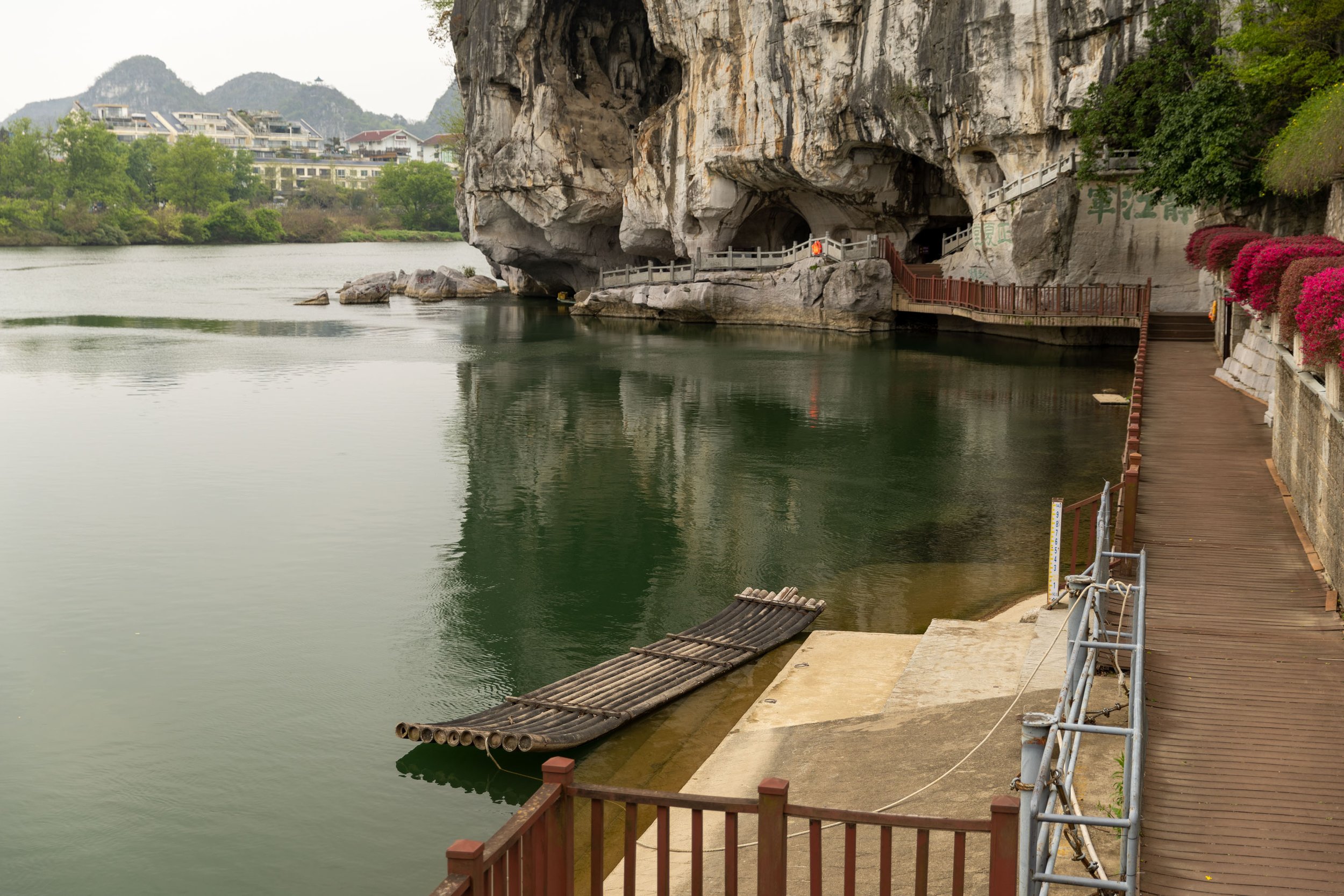 Riverbank with bamboo raft, wooden walkway, and rocky cave entrance in a scenic landscape.
