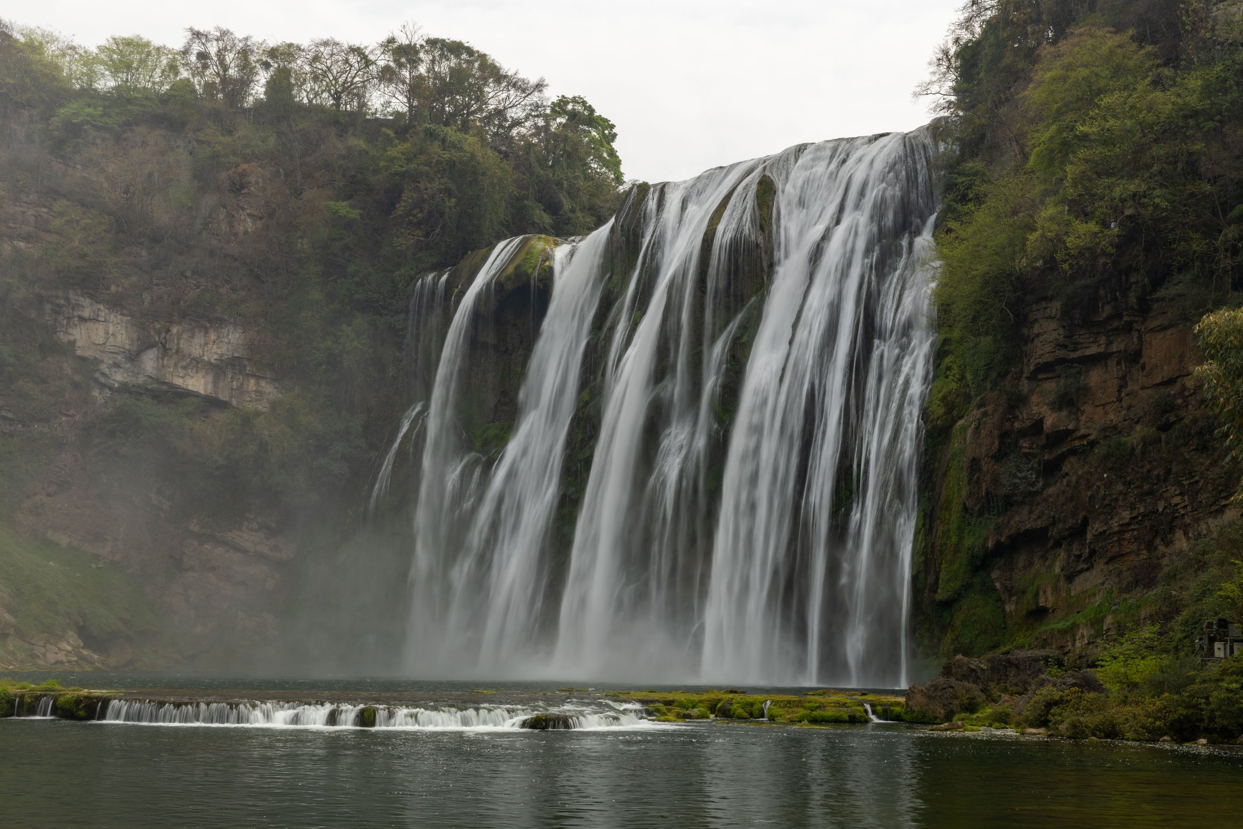 Waterfall cascading over rocks surrounded by lush greenery, with a pool of water at the base.