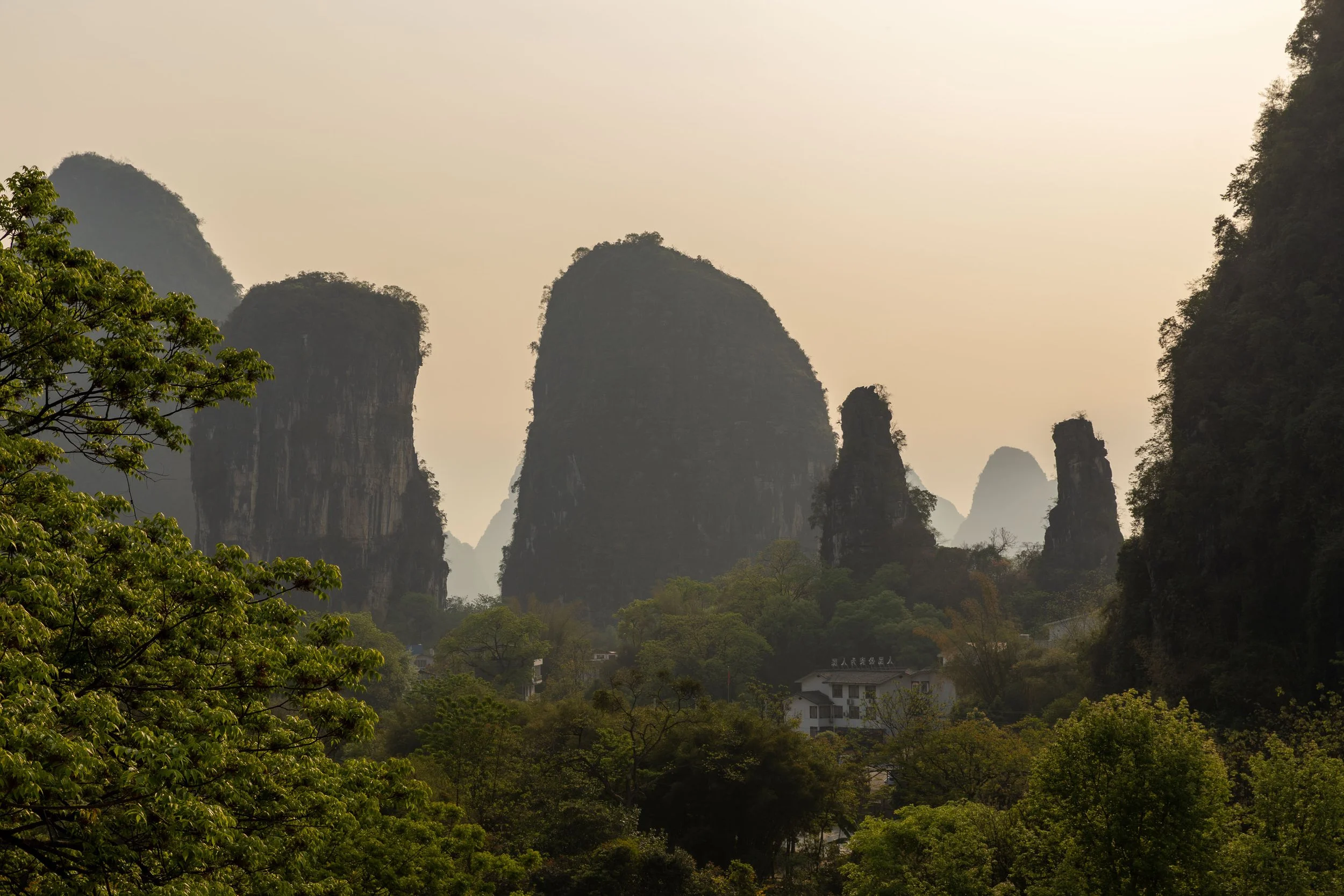 Scenic view of karst mountains in Guilin, China, with lush green trees in the foreground and a hazy sky.
