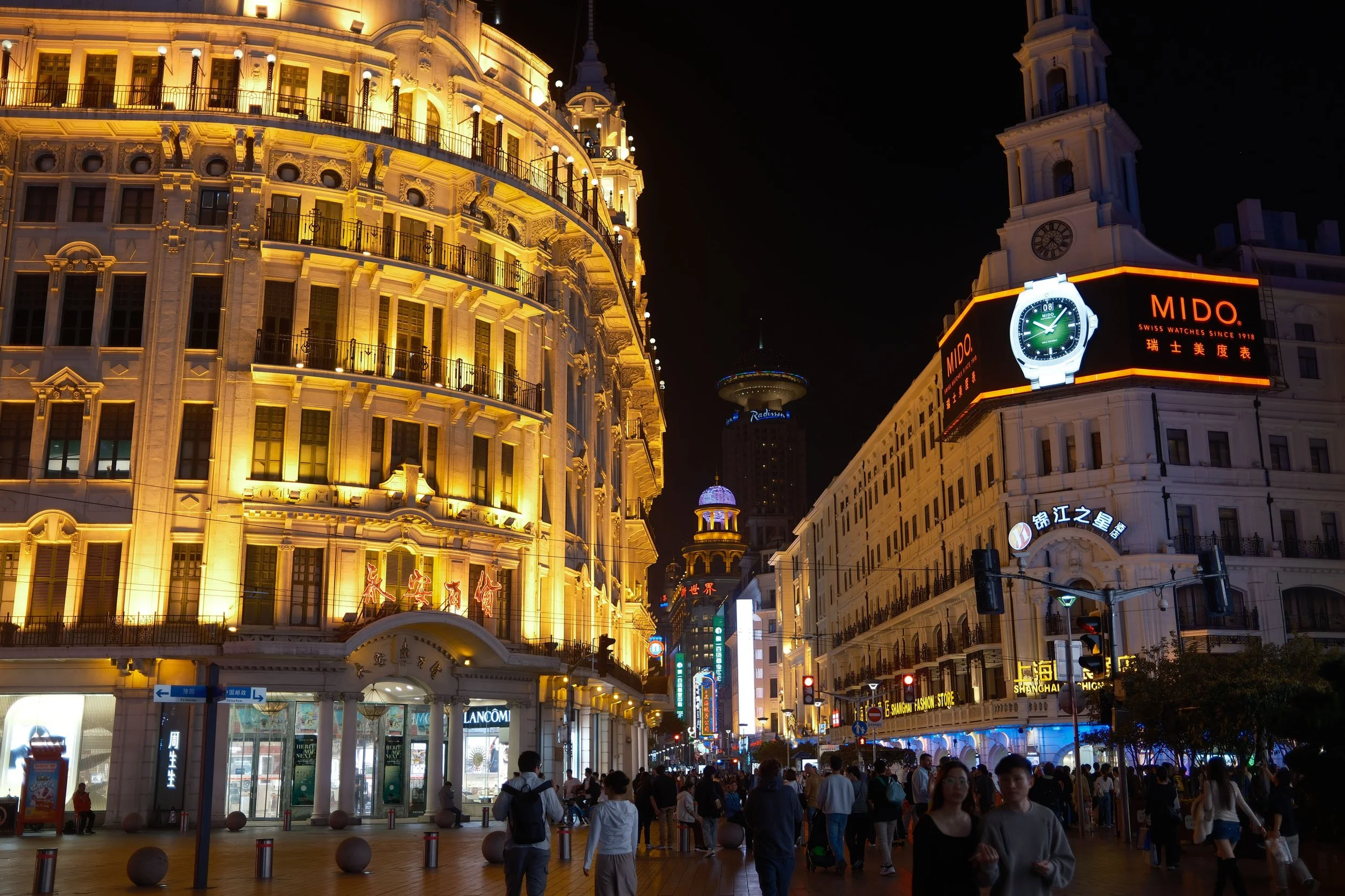 Night view of East Nanjing Road, Shanghai, with illuminated historic buildings and a crowded street, featuring the Mido watch billboard.